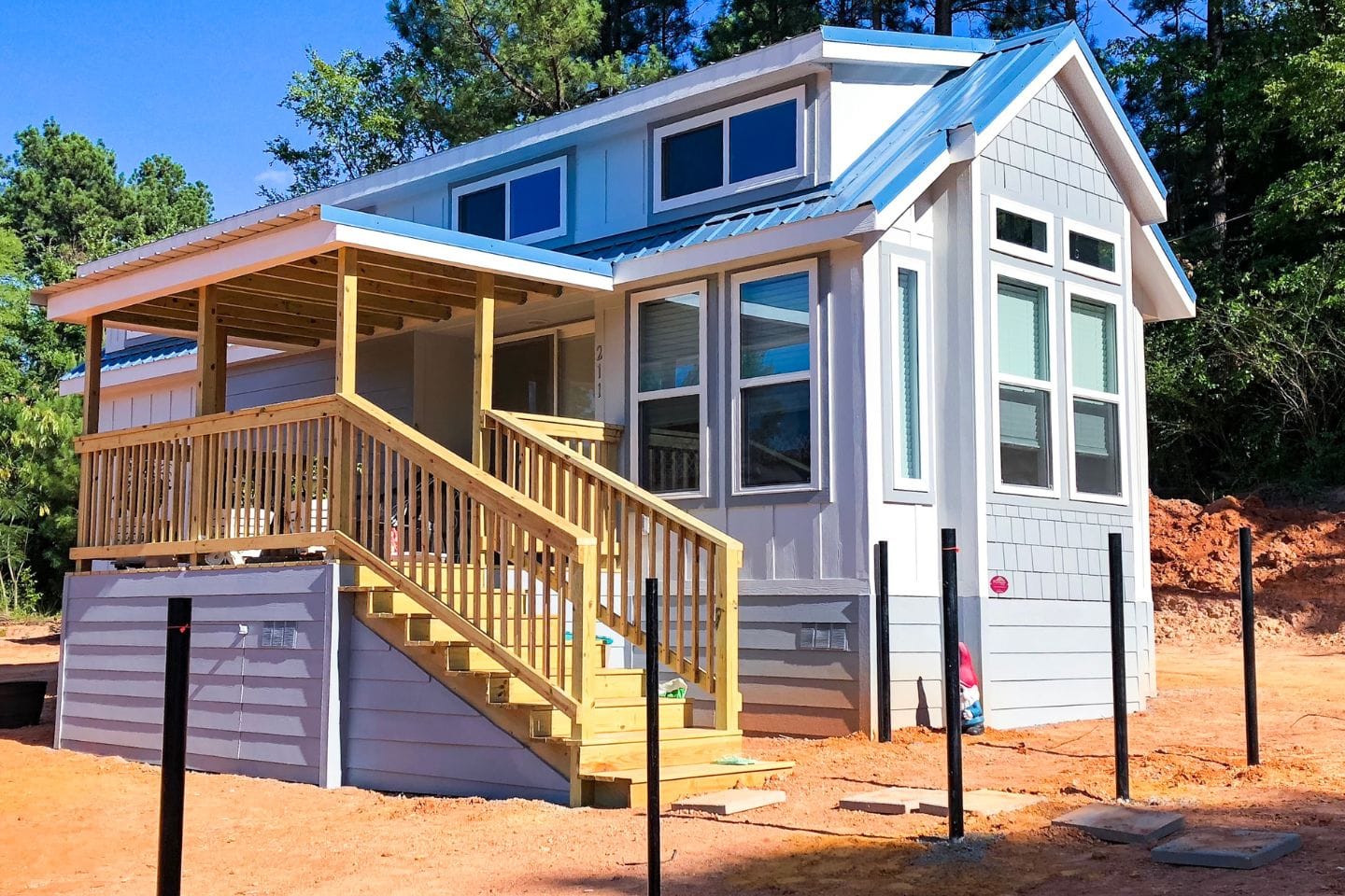 A small house with light gray siding, white trim, and a blue metal roof. It features large windows and a wooden porch with stairs. The area surrounding the house is under construction, possibly paving the way for future ADUs or secondary housing units.