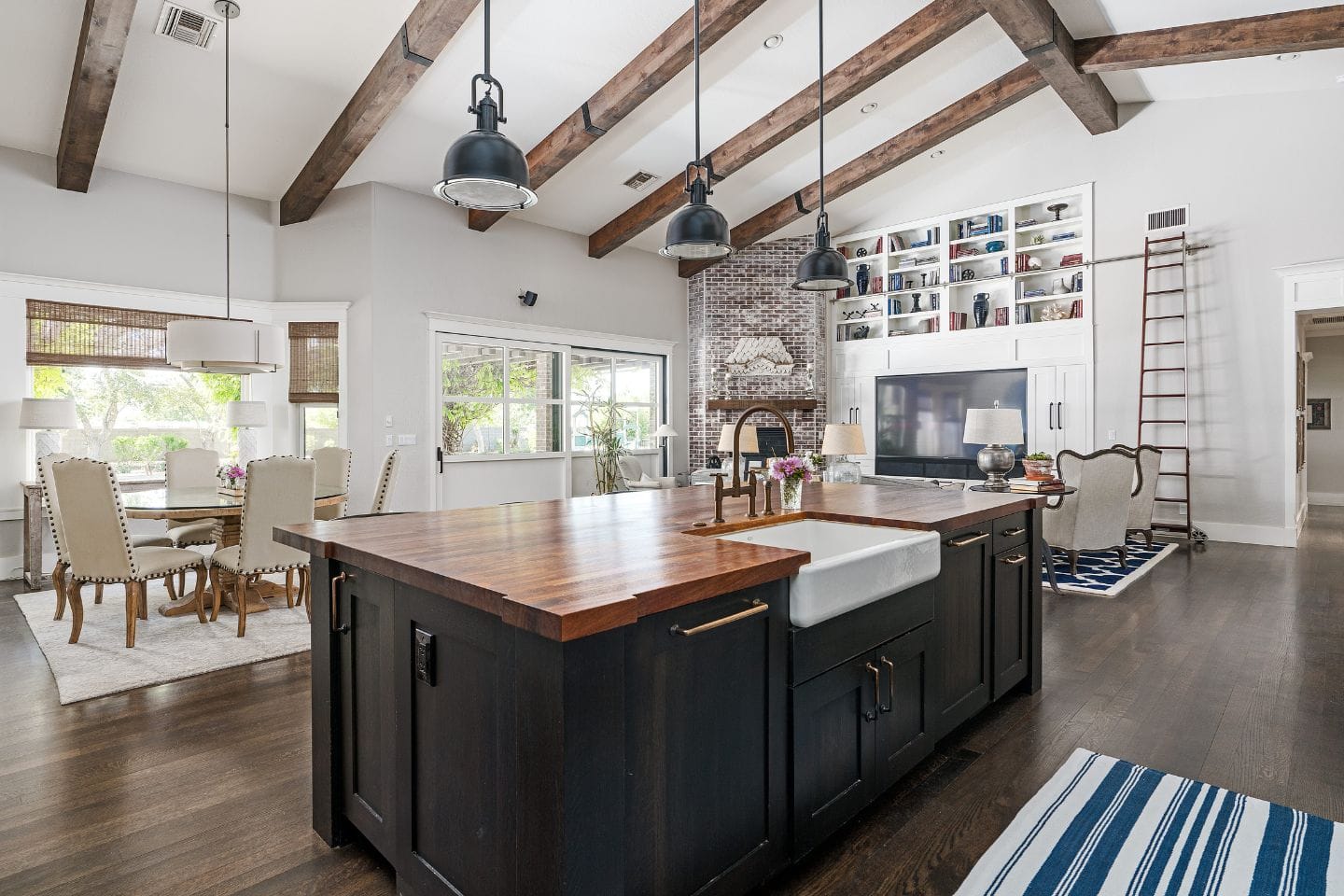 Open-concept home featuring a kitchen and living area with wooden beams, large island, farmhouse sink, dining table, brick fireplace, and built-in bookshelves with a ladder.