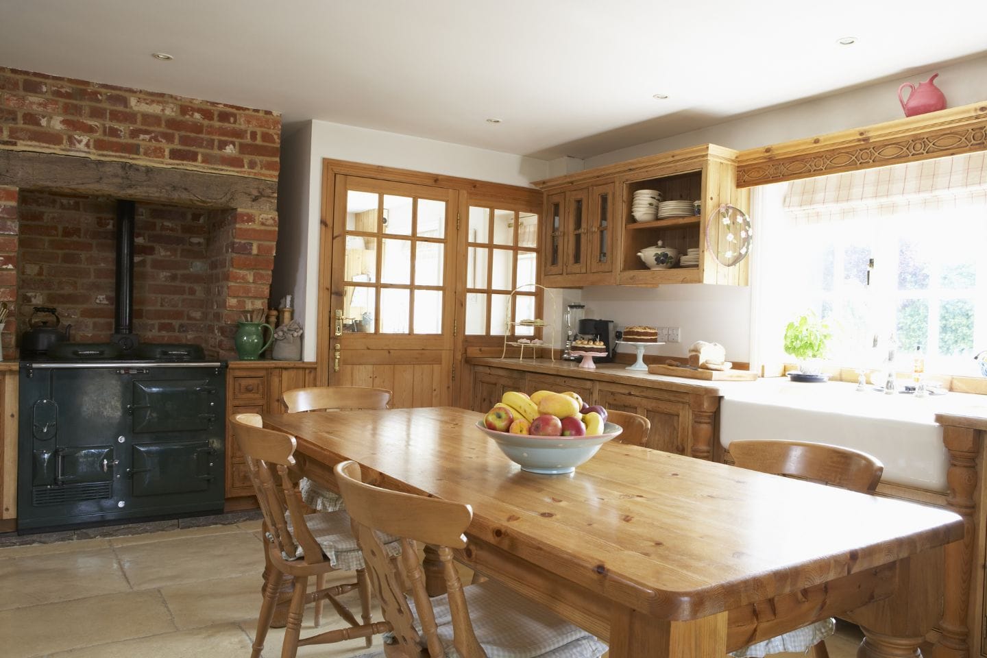 A rustic kitchen with a wooden dining table and chairs, a fruit bowl on the table, an exposed brick oven area, wood cabinets, and a large window letting in natural light, exudes the comforting warmth of home.