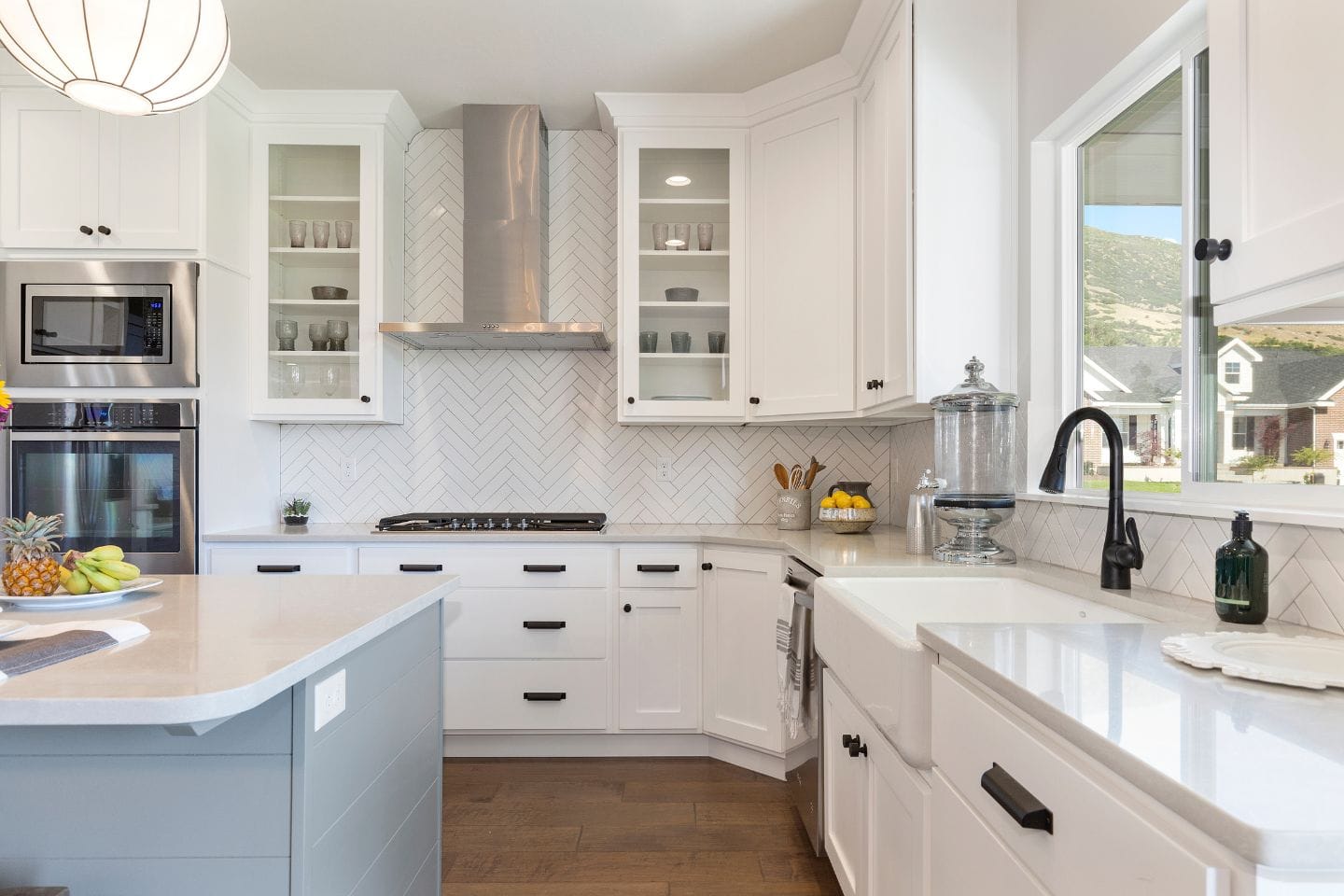 Modern home kitchen with white cabinets, stainless steel appliances, a farmhouse sink, and a central island. The backsplash features a herringbone tile pattern. A window offers a view of the outdoors.