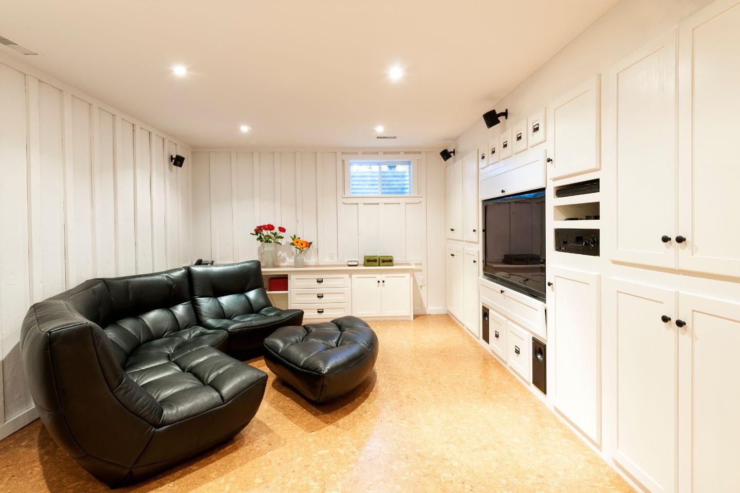 A cozy basement living room with a sectional leather sofa and ottoman, a wall-mounted TV, and built-in white cabinetry. Recent renovations include recessed lighting in the ceiling, enhancing the space where flowers on a counter add a touch of elegance.