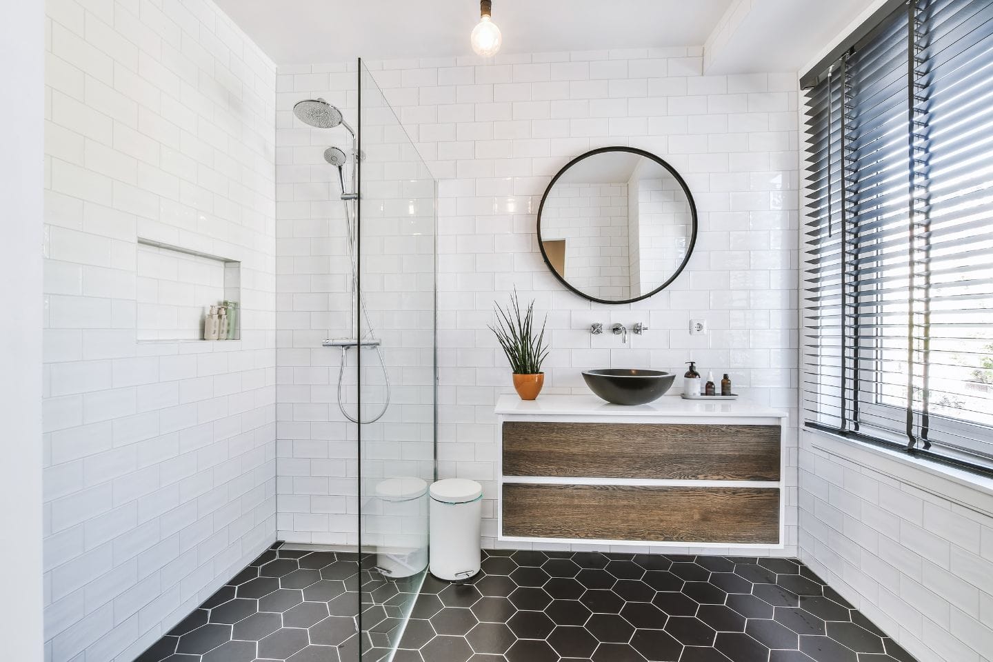 Modern bathroom with white subway tile walls, hexagonal black floor tiles, a round mirror above a vanity with a black sink, a walk-in shower with a glass partition, and a window with black blinds; perfect for any bath renovation.