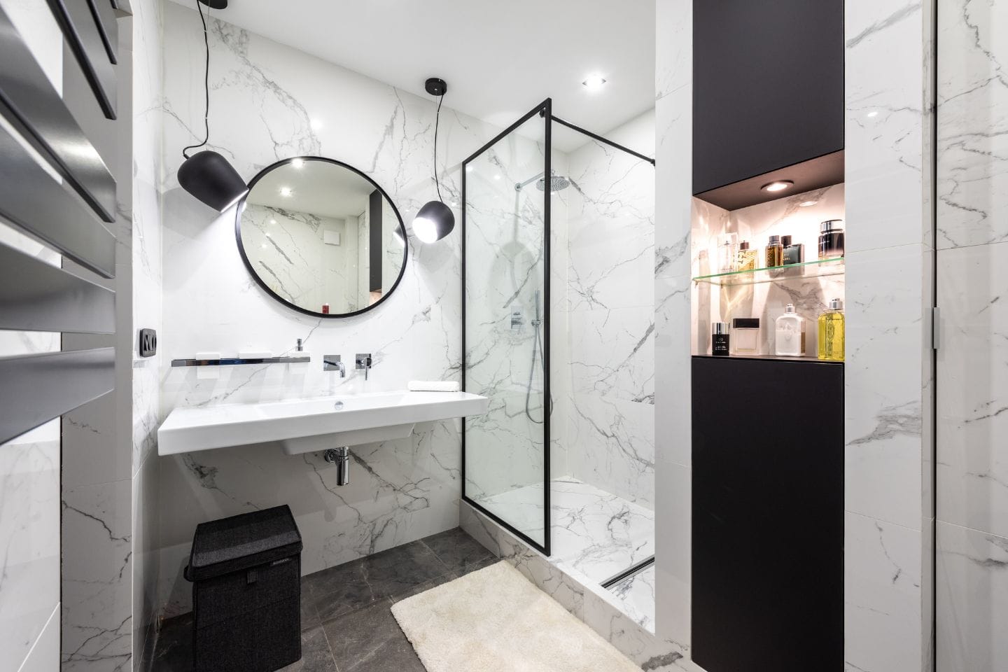 Modern bathroom with a marble design featuring a round mirror, wall-mounted sink, glass shower enclosure, and open shelving with toiletries. Perfect for anyone considering a tub to shower conversion during their bathroom remodeling.