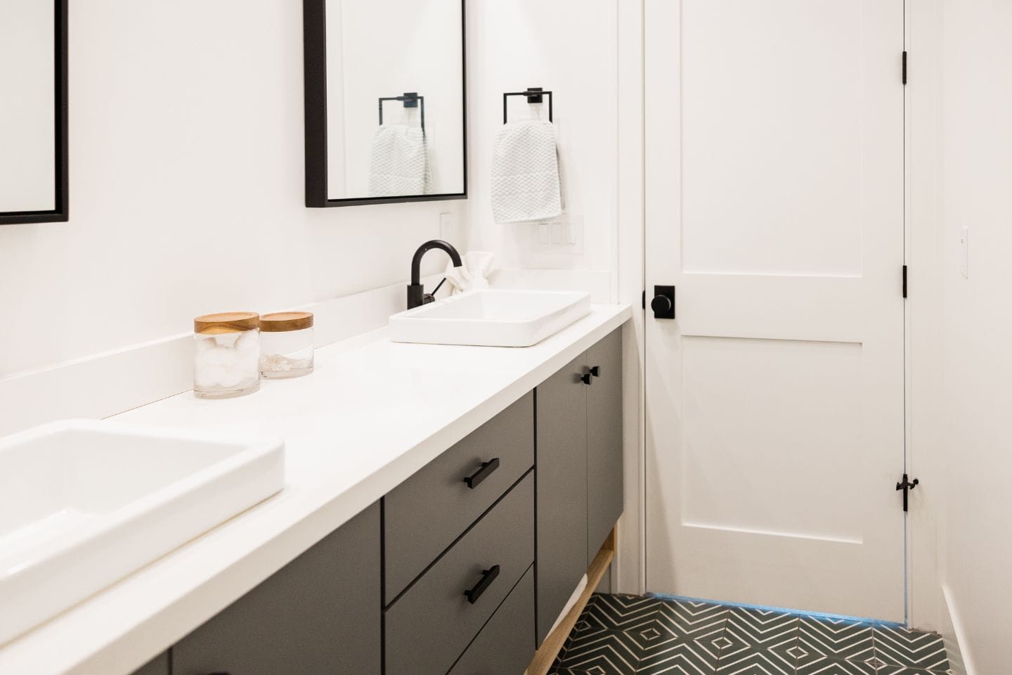 A modern bathroom with a white countertop, two vessel sinks, black fixtures, and gray cabinets. There are two mirrors above the sinks and a geometric-patterned floor, making it an ideal example of sleek bathroom renovations.