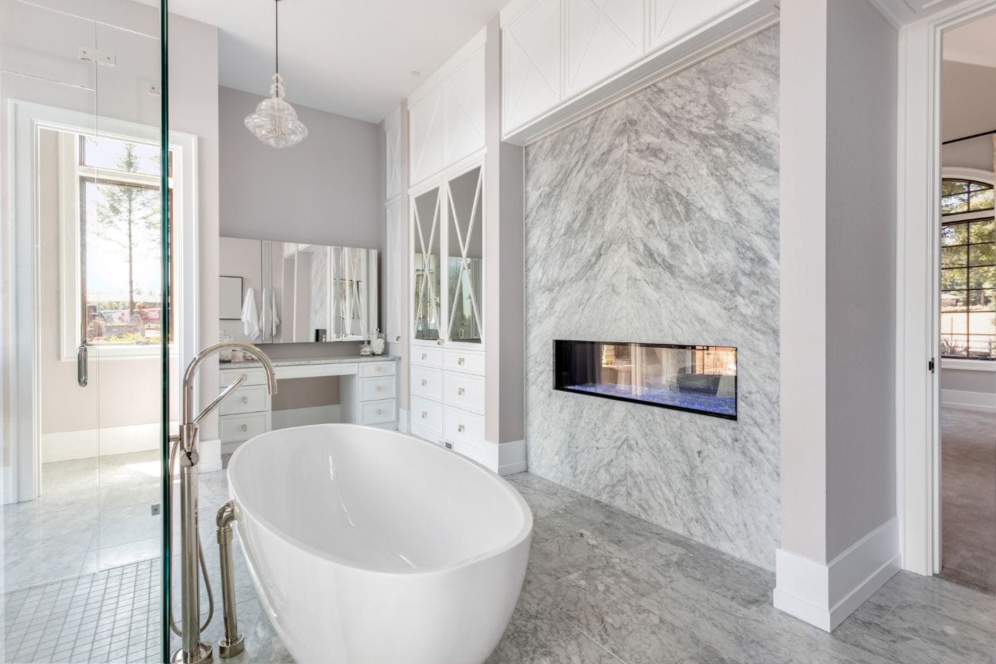 Modern bathroom, perfect for a tub to shower conversion, with a freestanding white bathtub, glass-enclosed shower, large mirror over a vanity, and a wall-mounted fireplace on a marble wall. Natural light streams in through the windows.