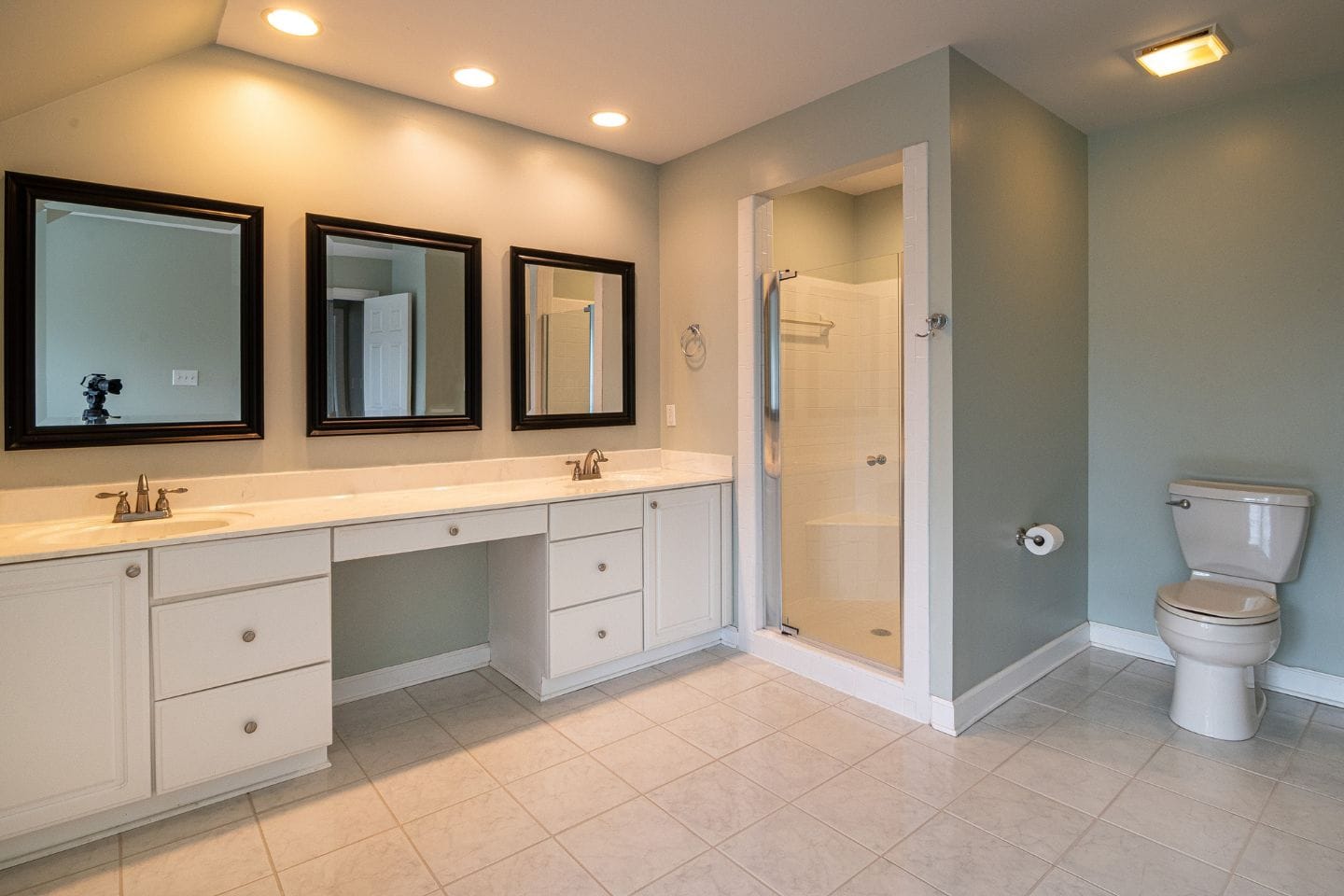 The bathroom features dual sinks and mirrors, a glass shower enclosure, and a toilet. With white cabinetry, grey walls, and beige tiled flooring, this Bath Conversions design offers both elegance and functionality.