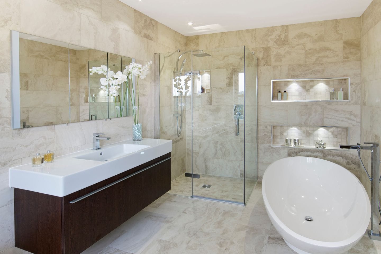 Modern bathroom with beige tiles, large mirror above a long sink counter, glass-enclosed shower from a recent tub to shower conversion, freestanding bathtub, and a vase of white flowers on the counter. Shelves with lights are visible.