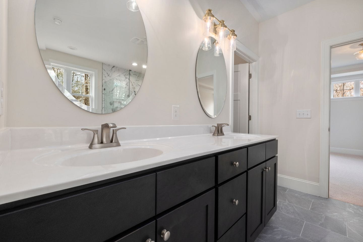 Modern bathroom with a dual-sink vanity, marble countertop, large oval mirrors, brushed nickel faucets, and wall-mounted light fixtures. The floor is tiled in gray, and natural light filters in from a window. A recent bath conversion replaced the tub with a sleek walk-in shower for added luxury.