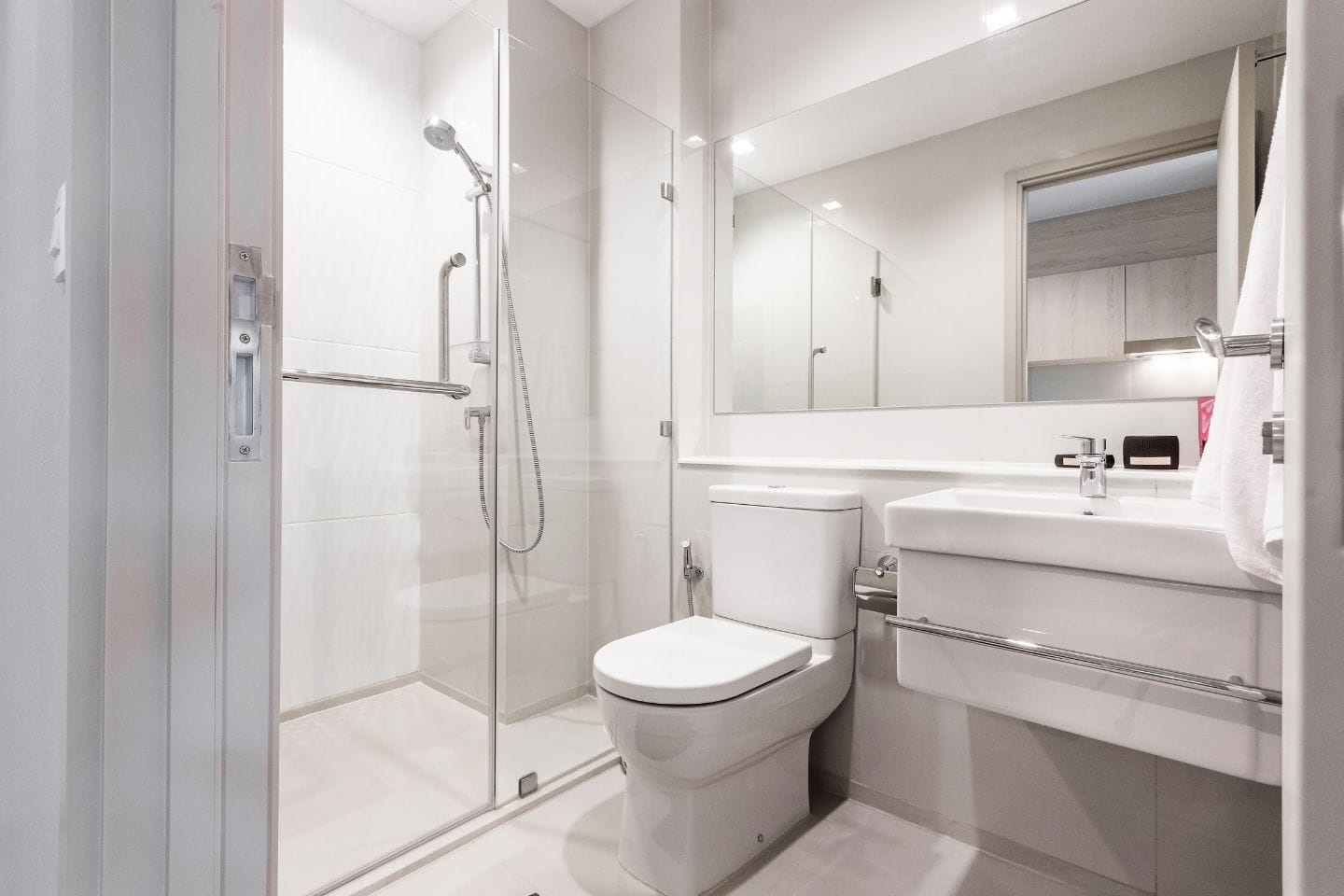 A modern bathroom features a glass-enclosed shower, a toilet, and a wall-mounted sink with a mirror above it. Bath conversions have enhanced this space, complete with a towel hanging on a rail.