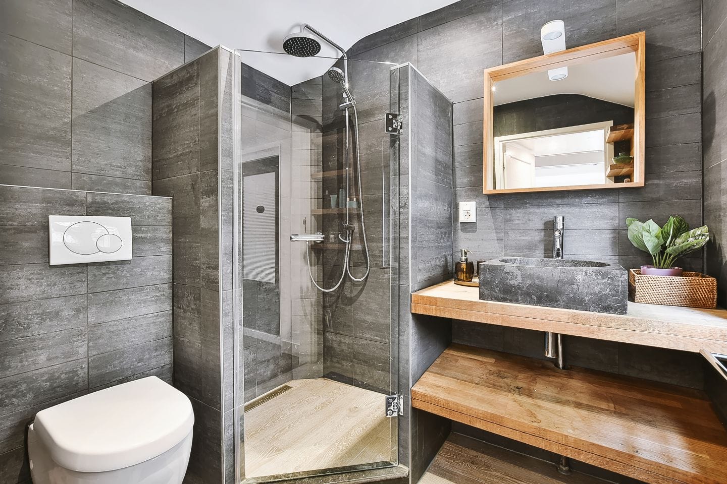 Modern bathroom with a glass-enclosed shower, square toilet, wood and stone sink vanity, large mirror, and grey tiled walls. Perfect for those considering a stylish bath renovation.