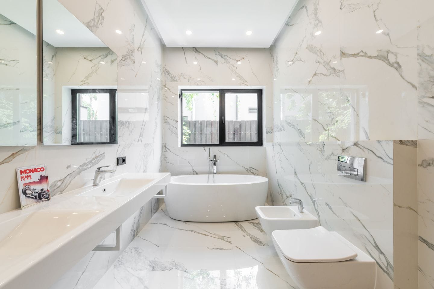 A modern bathroom renovation features a freestanding bathtub, a large mirror above a double sink, a wall-mounted toilet, and a bidet. Walls and floors are finished in white marble with gray veins.