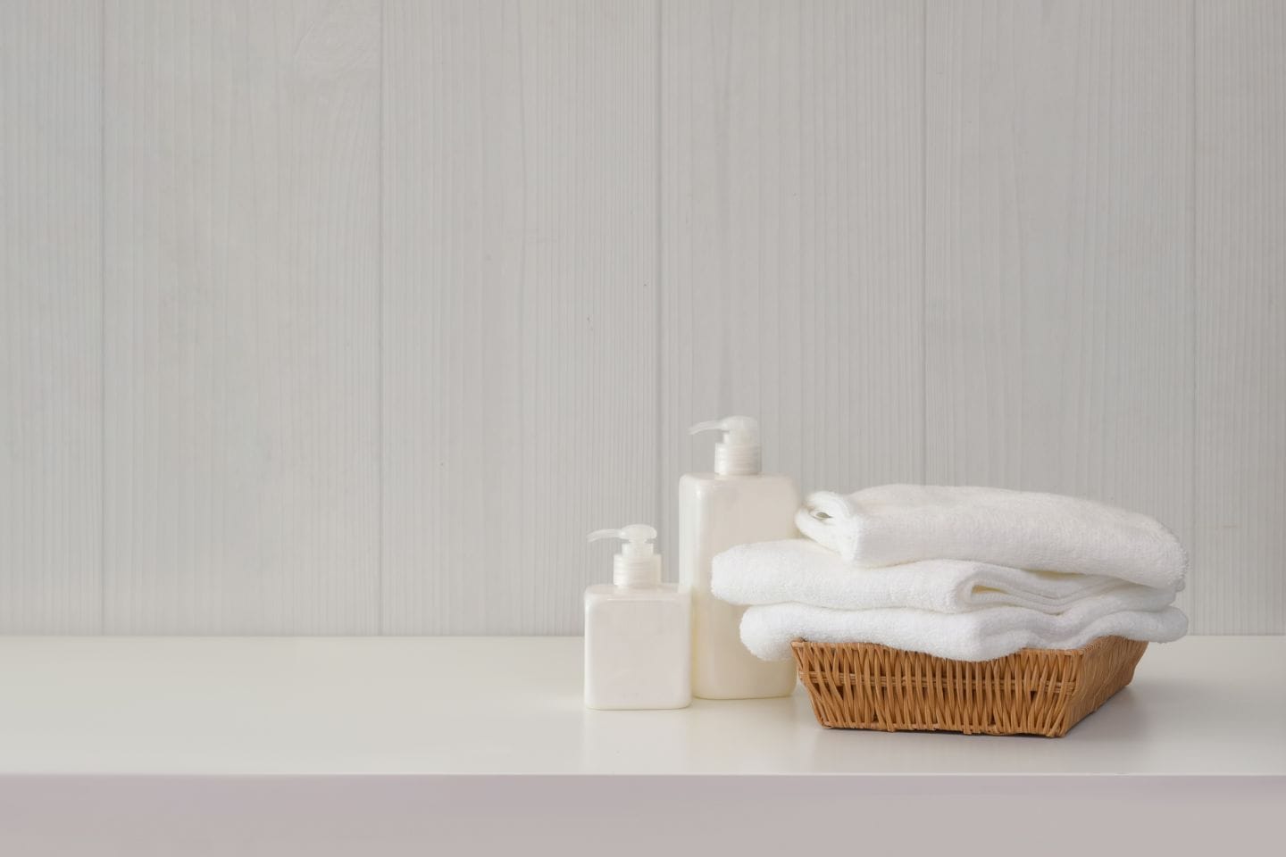 A wicker basket filled with folded white towels sits on a white surface alongside two white pump bottles, perfect for any bath conversion, against a light-colored wooden wall.