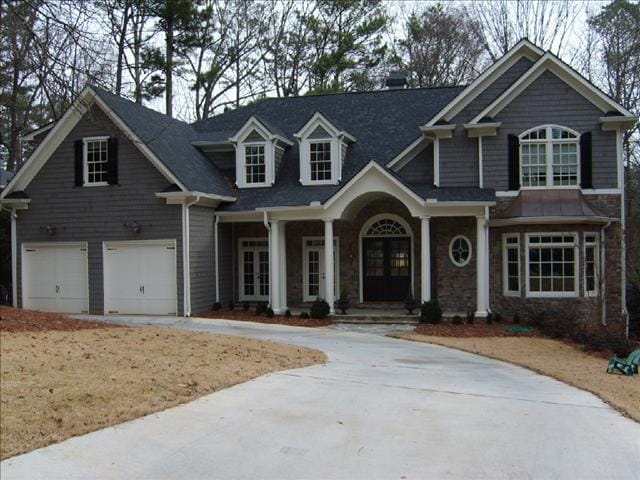 A two-story house with gray siding, white trim, and a dark shingled roof. It features a double garage, a front porch with columns, and several windows on both levels. The property is surrounded by trees and offers potential for room expansions to accommodate growing families.