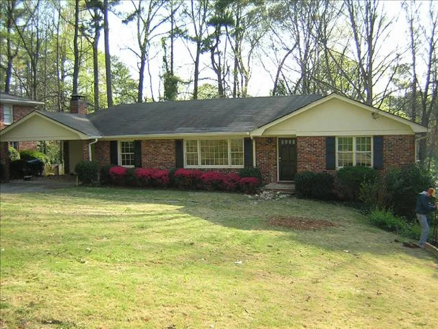 Single-story brick house with a gable roof, front porch, and large windows. Front yard with trimmed bushes, red azaleas, and a sloping lawn. A person is seen near the right edge of the image, possibly planning home expansions to enhance their charming residence.