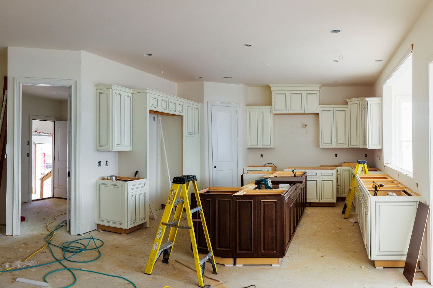 A kitchen under renovation with light-colored cabinets and an island. Tools and ladders occupy the space as cabinet installation progresses. Walls are partially painted, and the floor is covered to protect from construction debris.