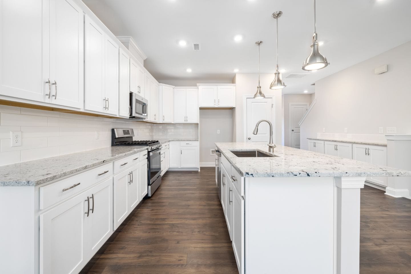 A modern kitchen featuring a seamless cabinet installation with white cabinets, stainless steel appliances, a large island with a sink, and wooden flooring. Three pendant lights hang above the island.