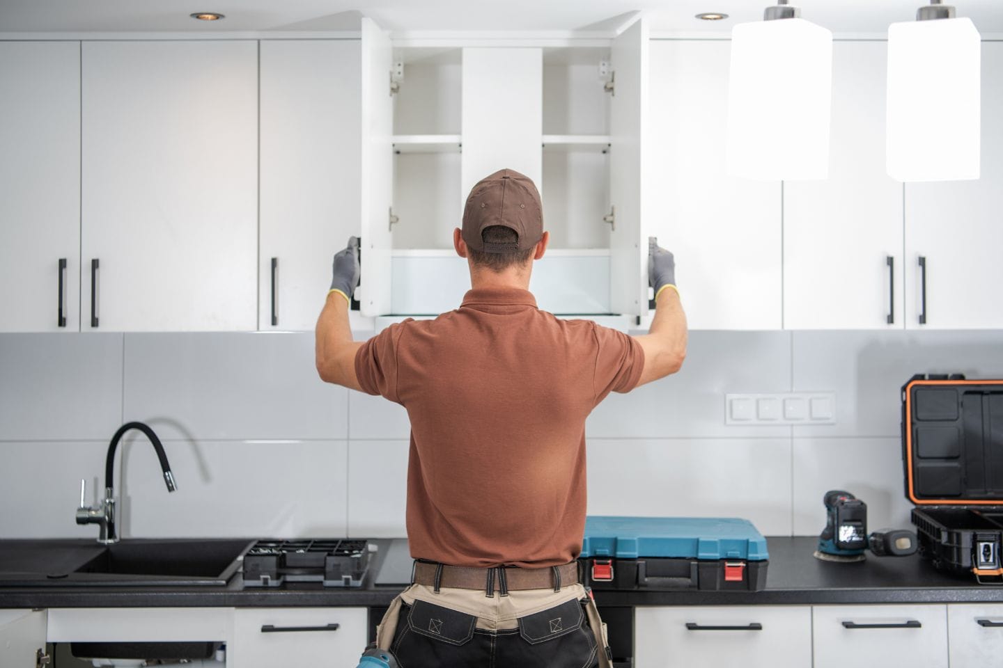 A person in a brown shirt and cap performs a cabinet installation in a modern kitchen with black countertops and various tools scattered around.