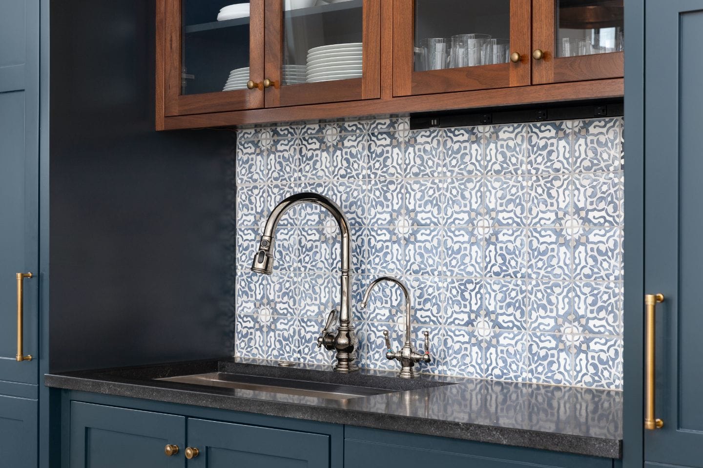 A kitchen sink installation features a brushed nickel faucet and a smaller faucet against a blue and white patterned backsplash, complemented by dark countertops and wooden cabinets.