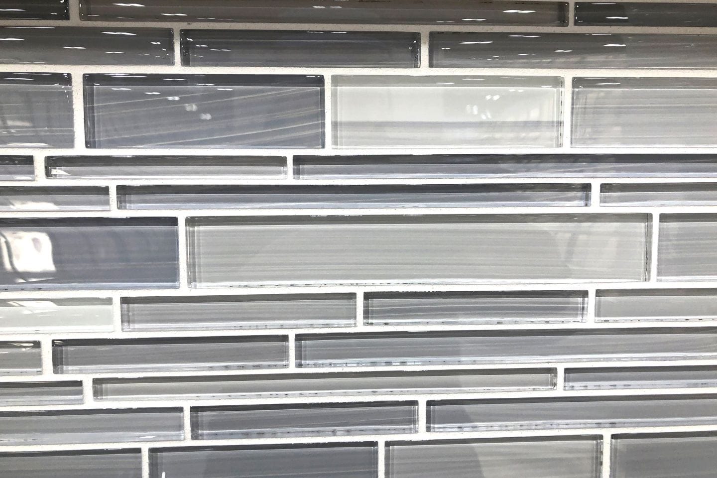 Close-up of a wall with a pattern of rectangular glass tiles in varying shades of gray and white. The backsplash installation features an offset design with white grout, perfect for any home improvement project.