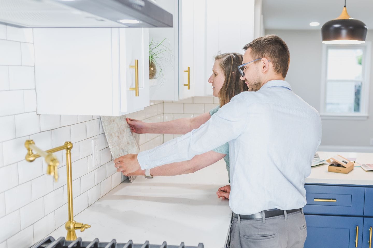 Two people inspect a tile sample against a white kitchen backsplash, standing by a counter with cabinets and a stove, discussing installation techniques for their latest home improvement project.