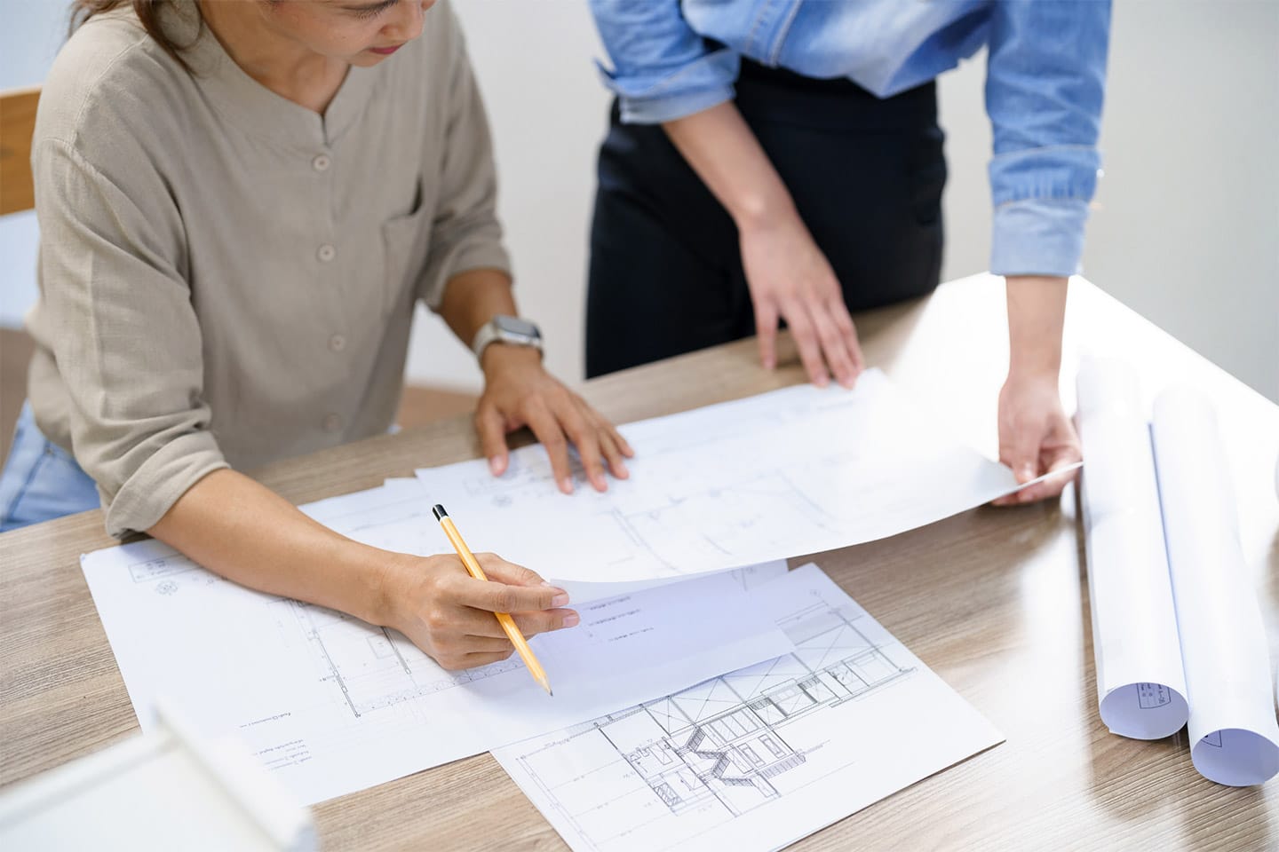 Two people at a desk reviewing home architectural blueprints; one person is seated and writing, while the other stands, pointing at the papers.