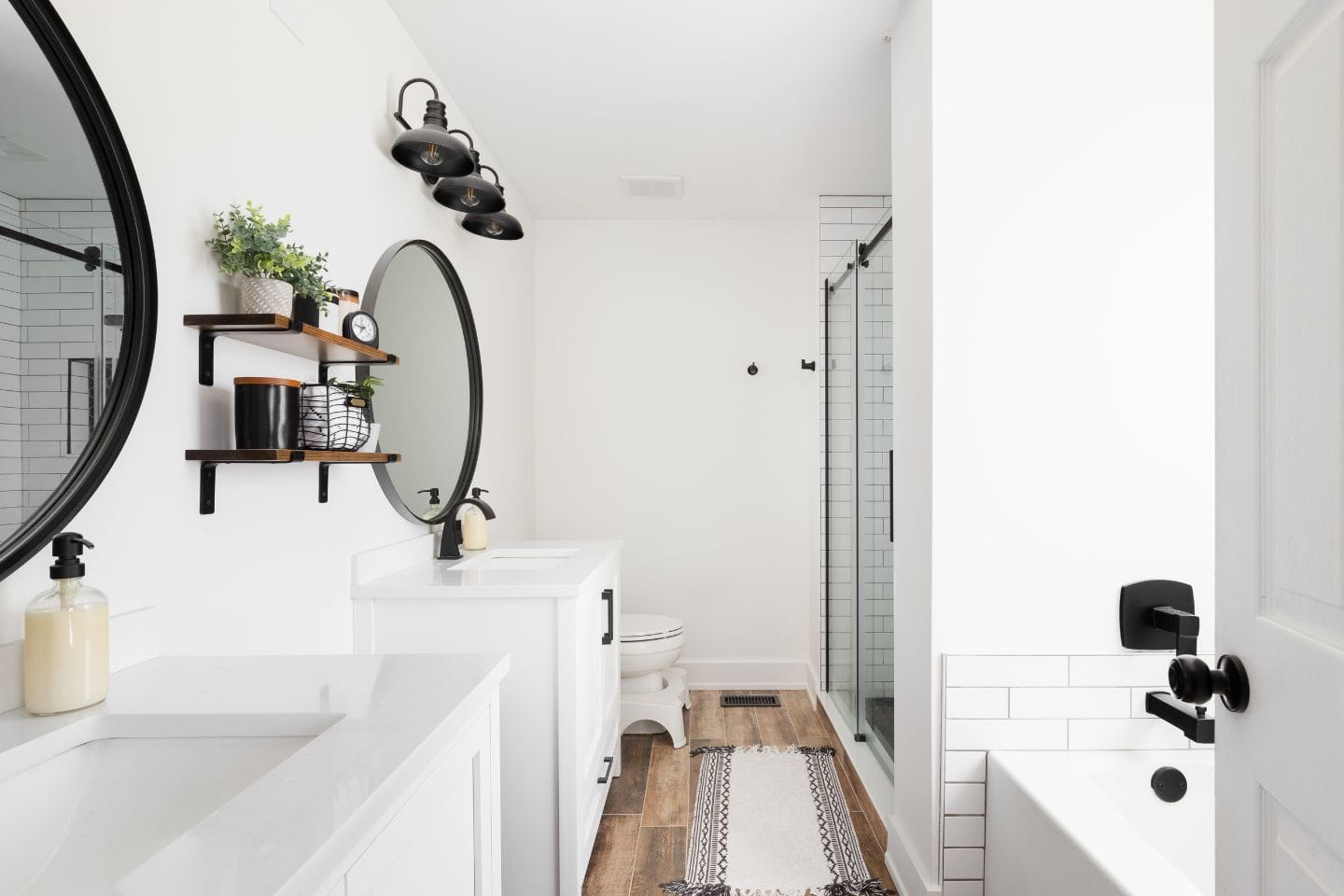 A modern bathroom with dual sinks, round mirrors, wall-mounted shelves, a walk-in shower with glass doors, white tiles, and a light wood floor. A black and white rug lies on the floor.