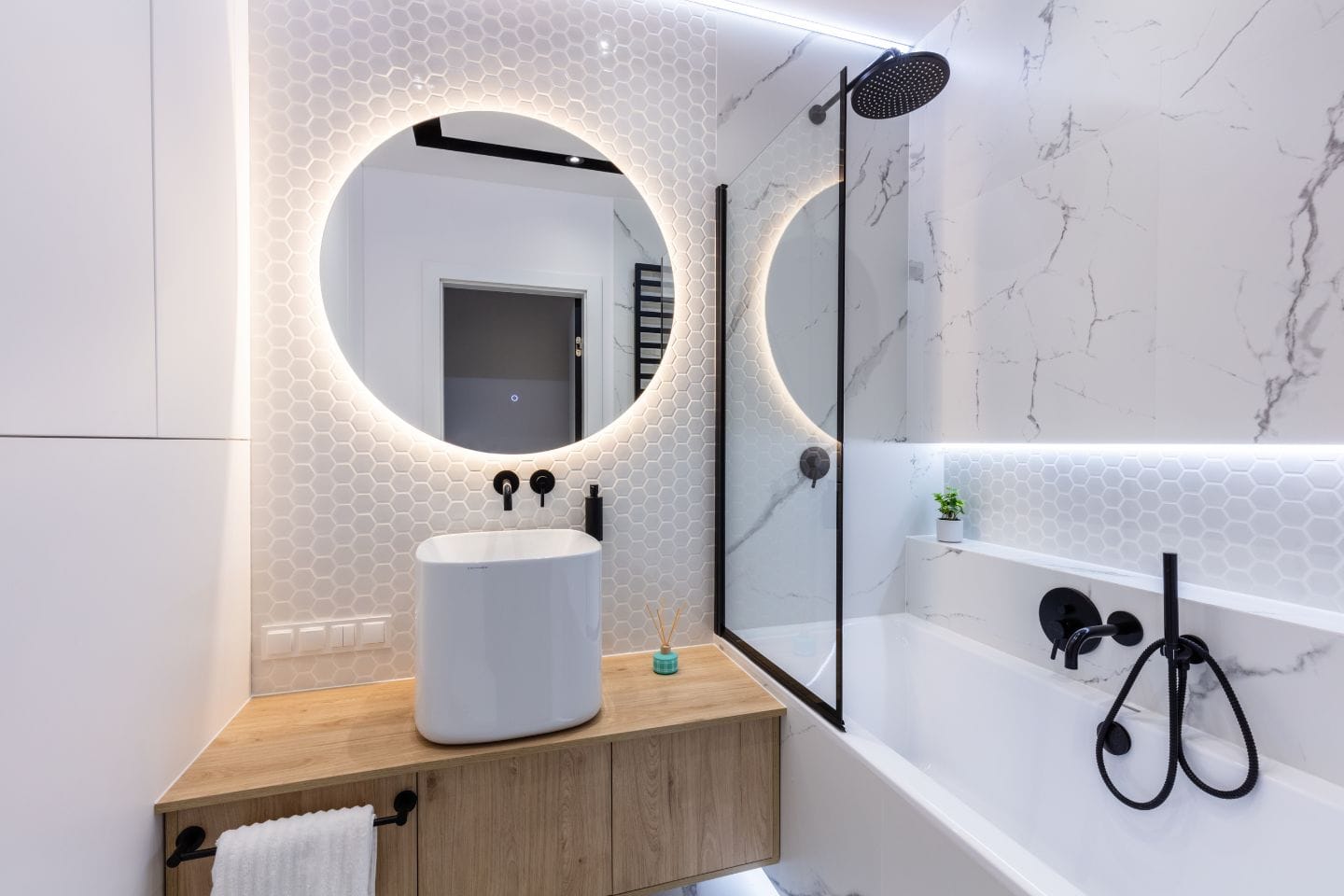 Modern bathroom with a backlit circular mirror, white hexagonal tile backsplash, a vessel sink, a black wall-mounted faucet, and a tub with a black shower fixture on a marble-patterned wall.