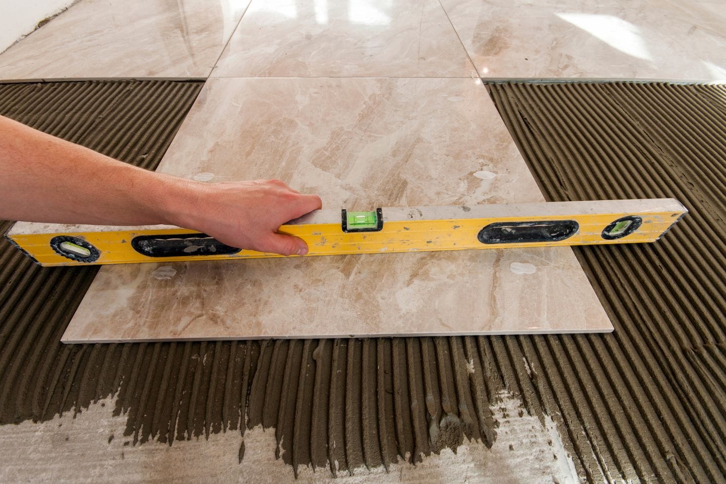 A person uses a yellow spirit level to ensure that a marble tile is being installed flat and evenly on adhesive mortar, achieving perfect tiling for the flooring project.