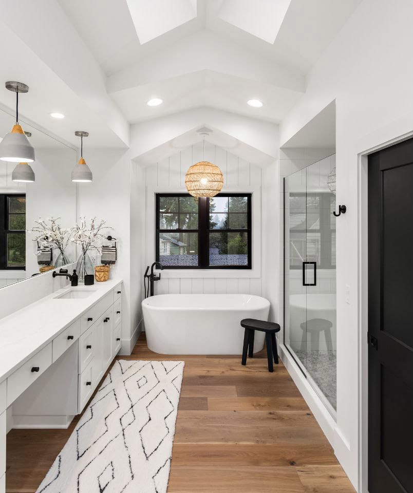 Modern bathroom with a freestanding tub, black-framed window, and glass-enclosed shower. Features white cabinetry, a large mirror, and hardwood flooring, accented by a patterned rug and pendant lights.