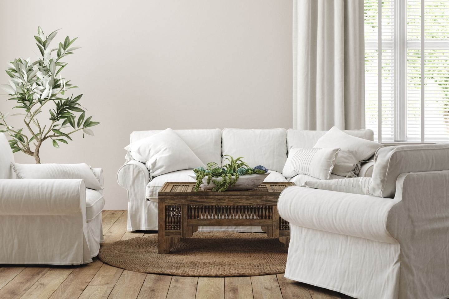 A living room with white upholstered furniture, a wooden coffee table, a potted plant, and a round woven rug on a wooden floor. White curtains hang by the window.