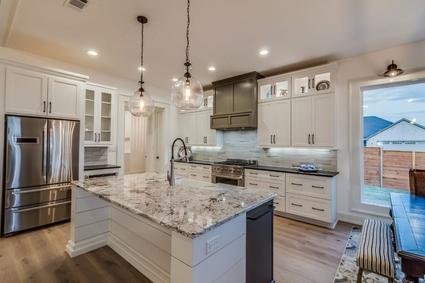 Modern kitchen with white cabinetry, stainless steel appliances, and a large marble island. The room is illuminated by pendant lights and has wooden flooring. A window provides natural light, highlighting the pristine kitchen surfaces and elegant marble counters.