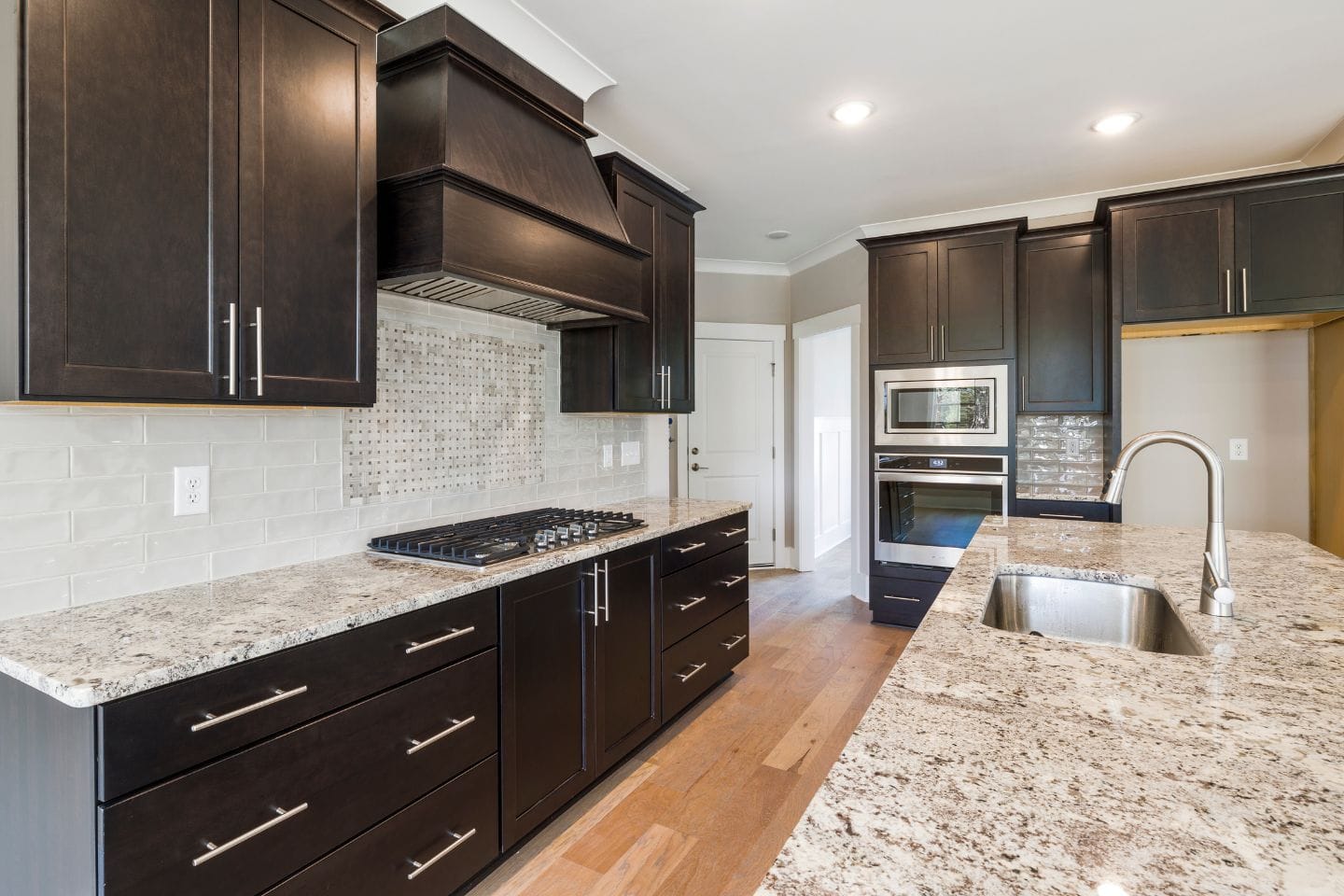 Modern kitchen with dark wood cabinets, granite countertops, stainless steel appliances, and a gas stovetop. Neutral backsplash tiles and recessed lighting enhance the clean, contemporary design of these exquisite kitchen countertops.