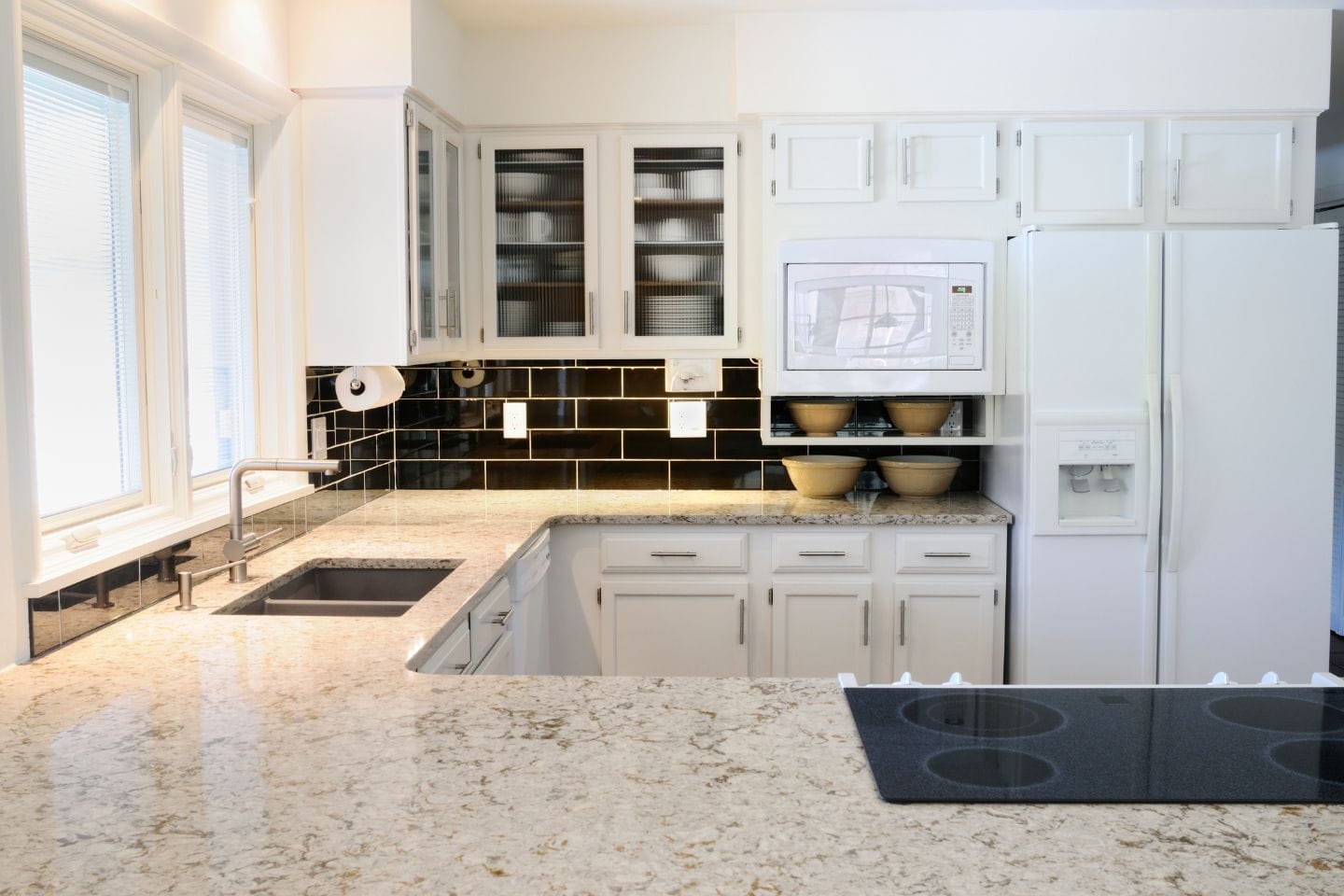 Modern kitchen with white cabinetry, black backsplash, granite countertops, and a built-in microwave. The sleek design includes a refrigerator and stovetop. Large windows flood the space with natural light, making this kitchen both functional and inviting.