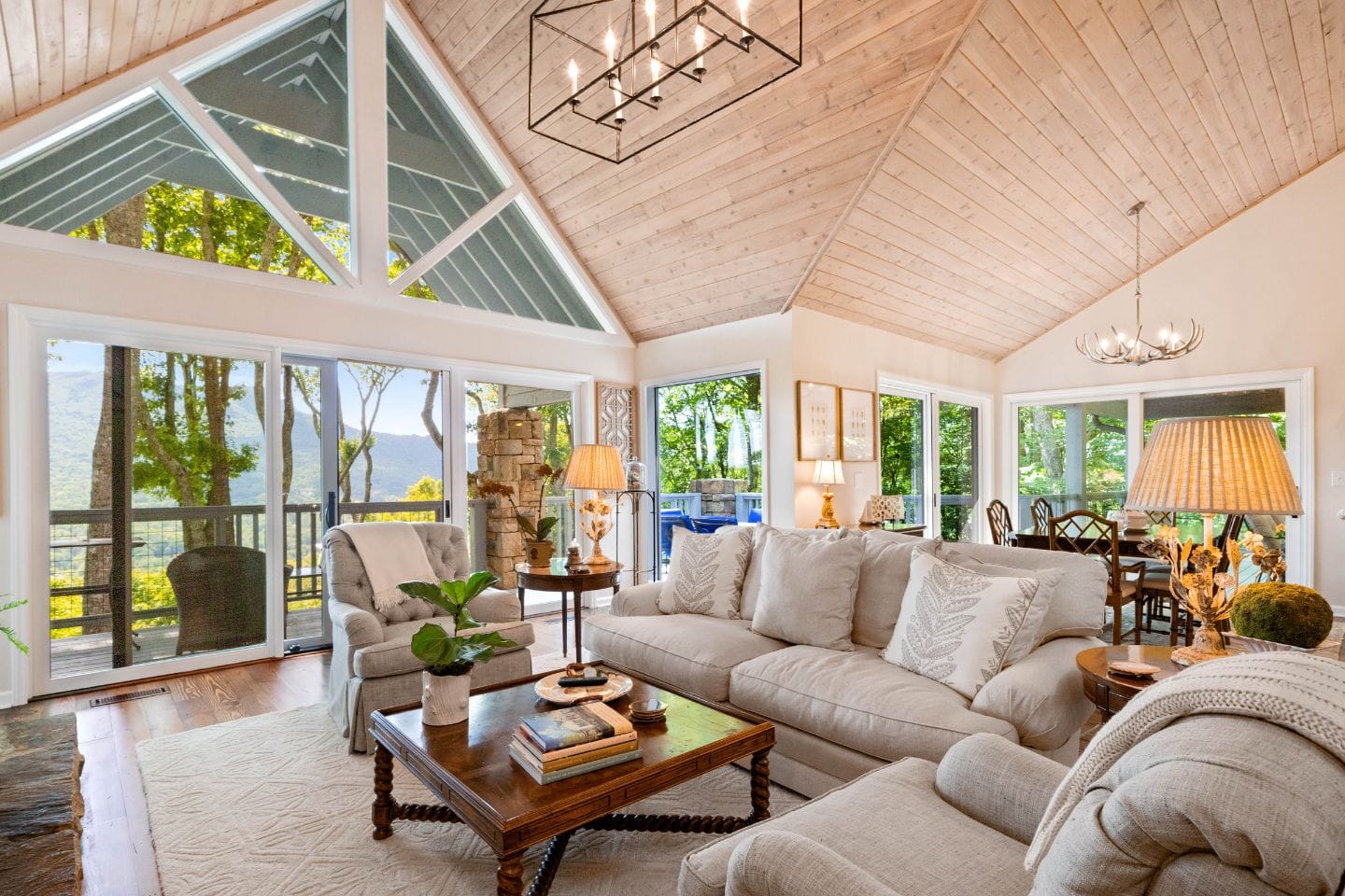 A cozy living room with large windows, light-colored couches, a wooden coffee table, and a vaulted ceiling. The view outside shows trees and a clear sky.