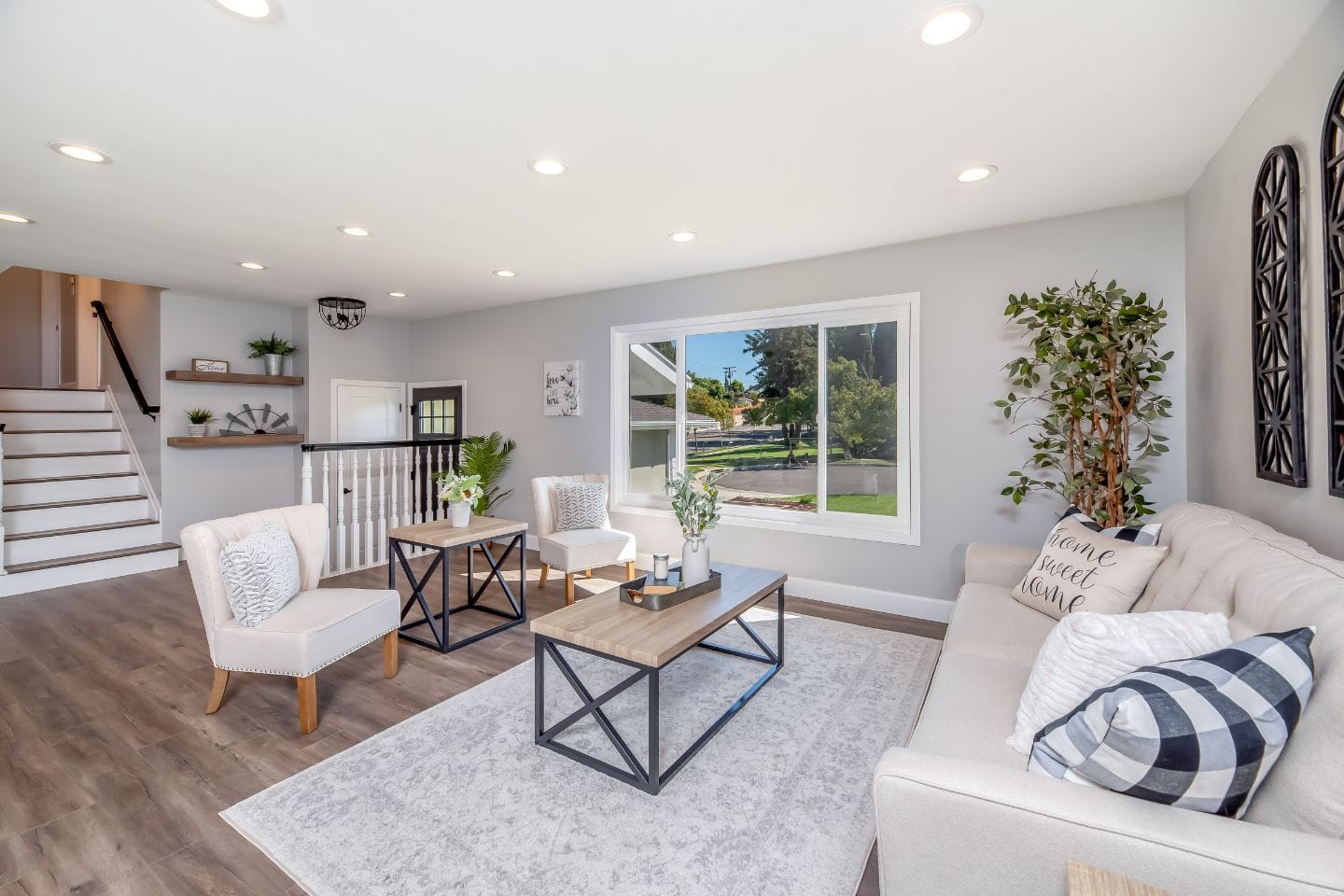 A modern living room with a beige sofa, two white armchairs, a light wood coffee table, and a large window overlooking a green yard. There are decorative pillows, plants, and wall art.