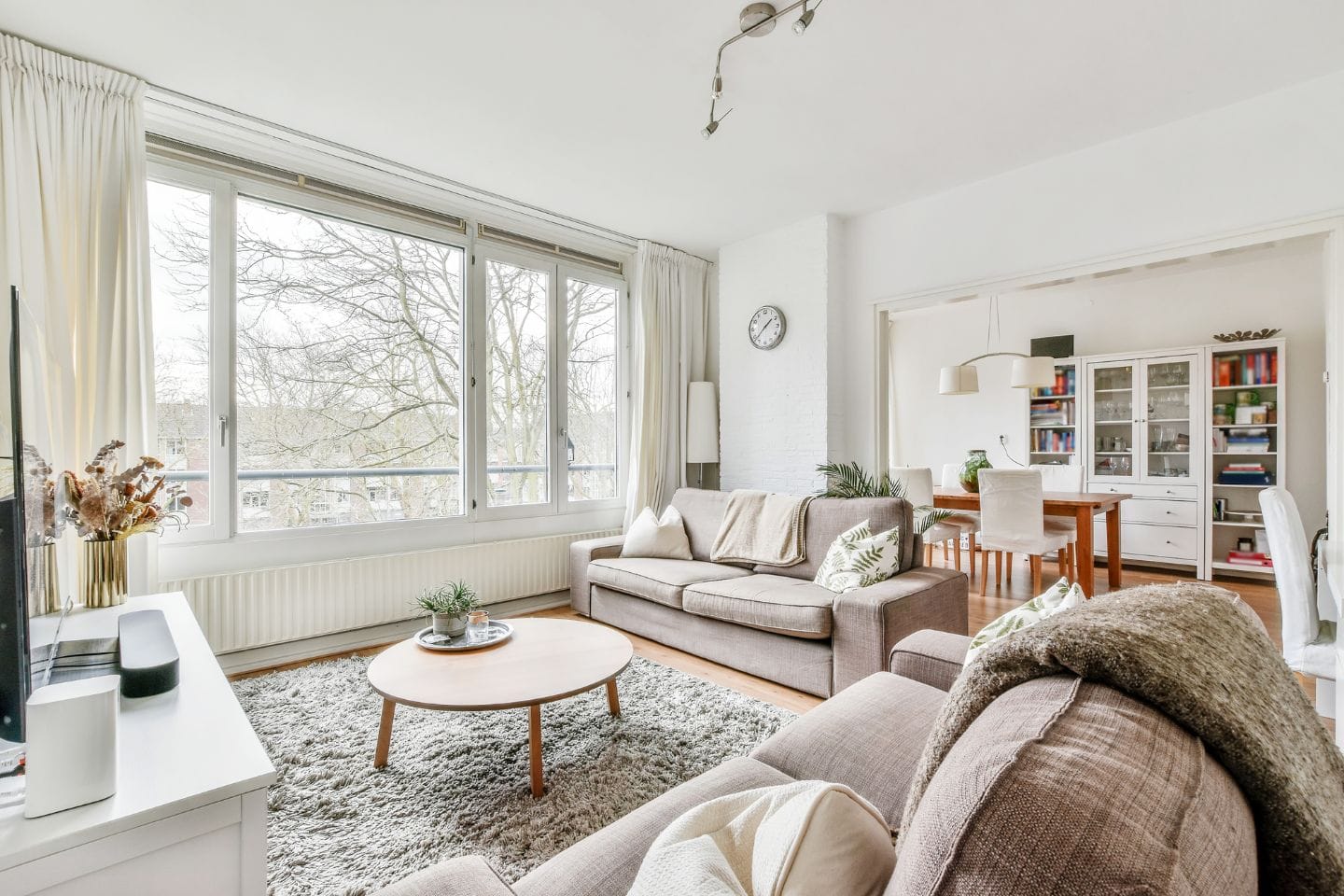 A bright living room with large windows, beige sofas, a round wooden coffee table, a rug, wall clock, and a dining area with a table, chairs, and a glass cabinet in the background.