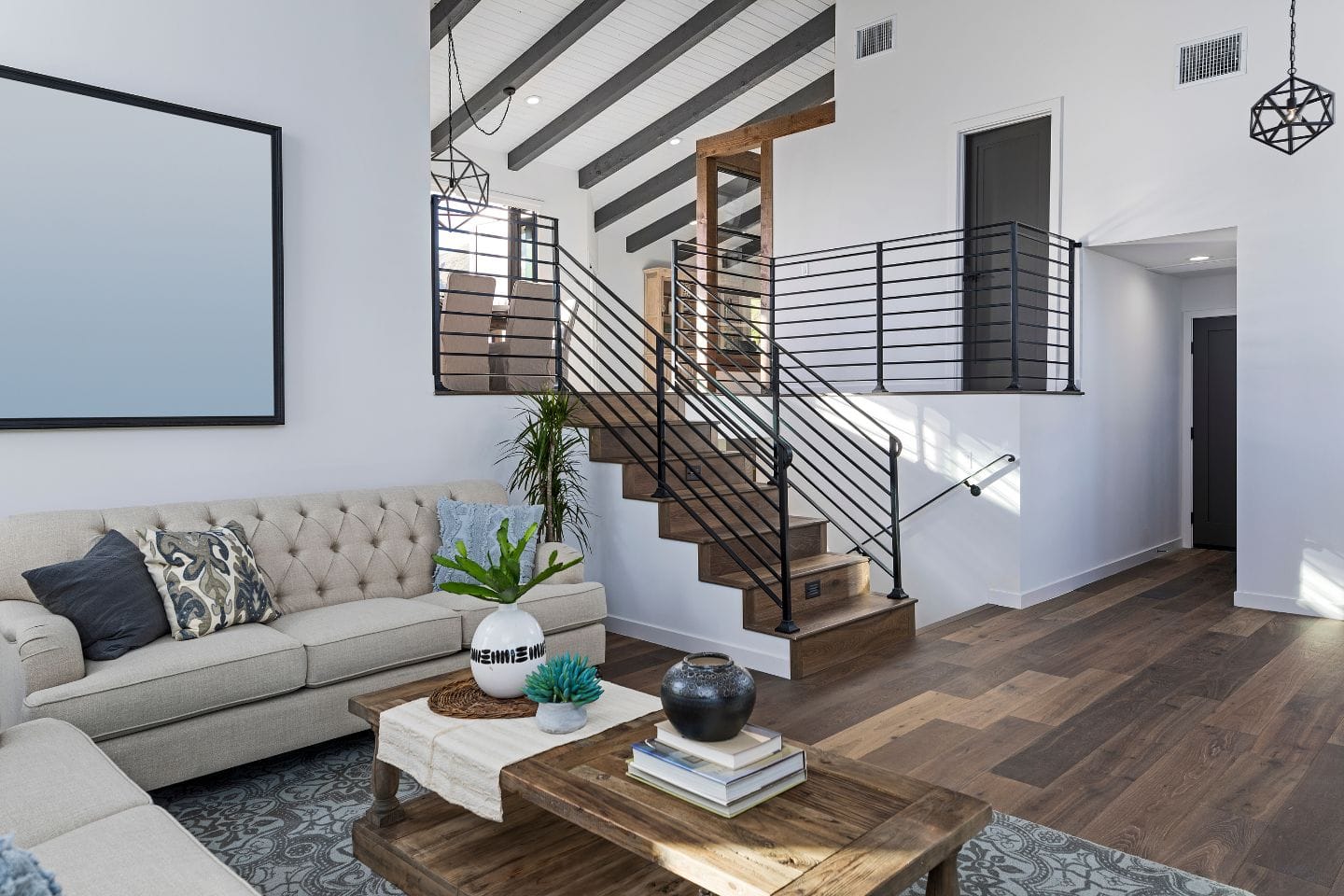 A modern living room with a beige sofa, wooden coffee table, staircase with black metal railings, hardwood floors, and high ceilings with exposed beams.