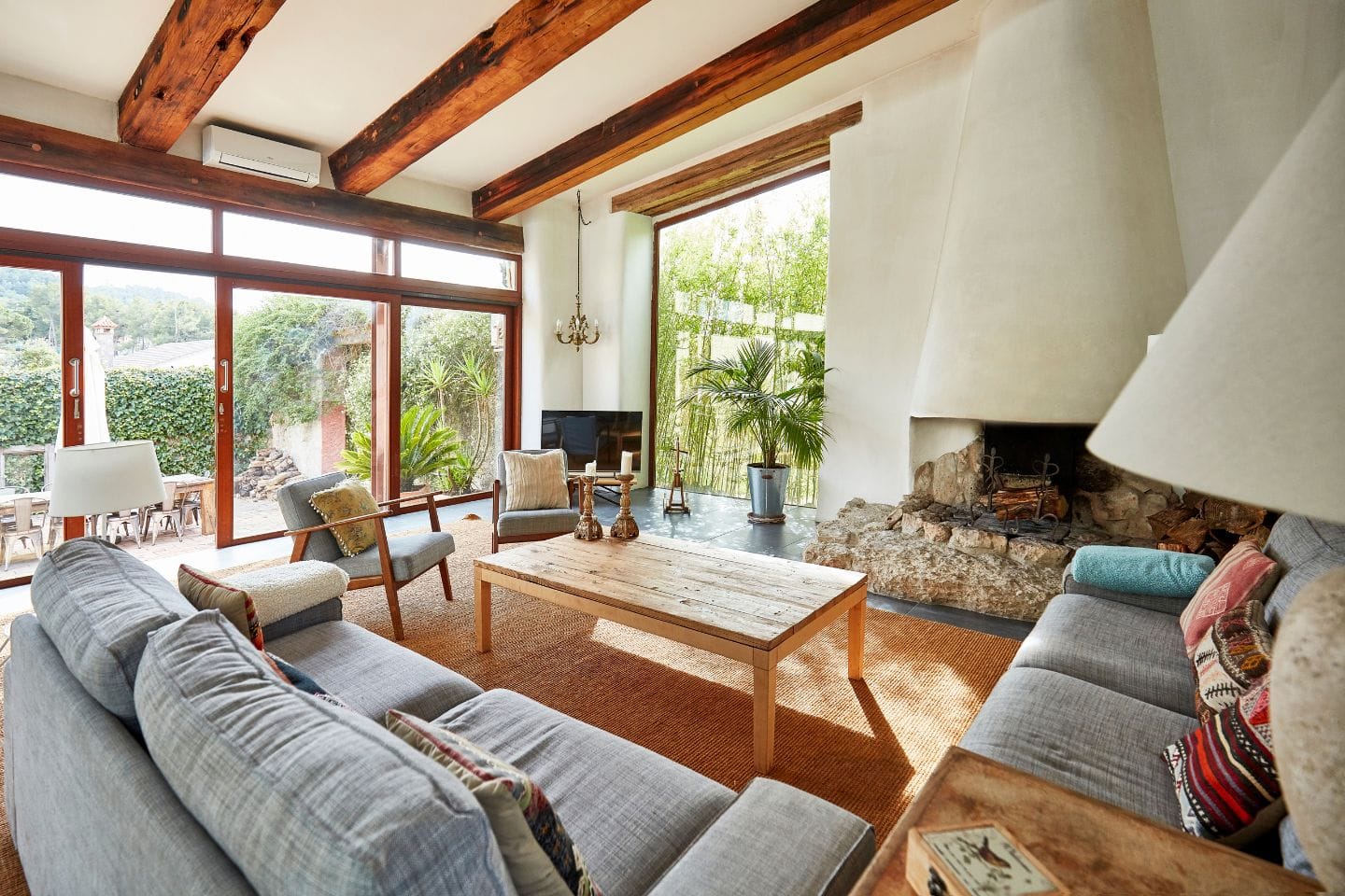 A living room with large windows, wooden beams, a light gray sofa, a wooden coffee table, and a corner fireplace. There's a patio visible through the open glass doors, and plants are placed around the room.