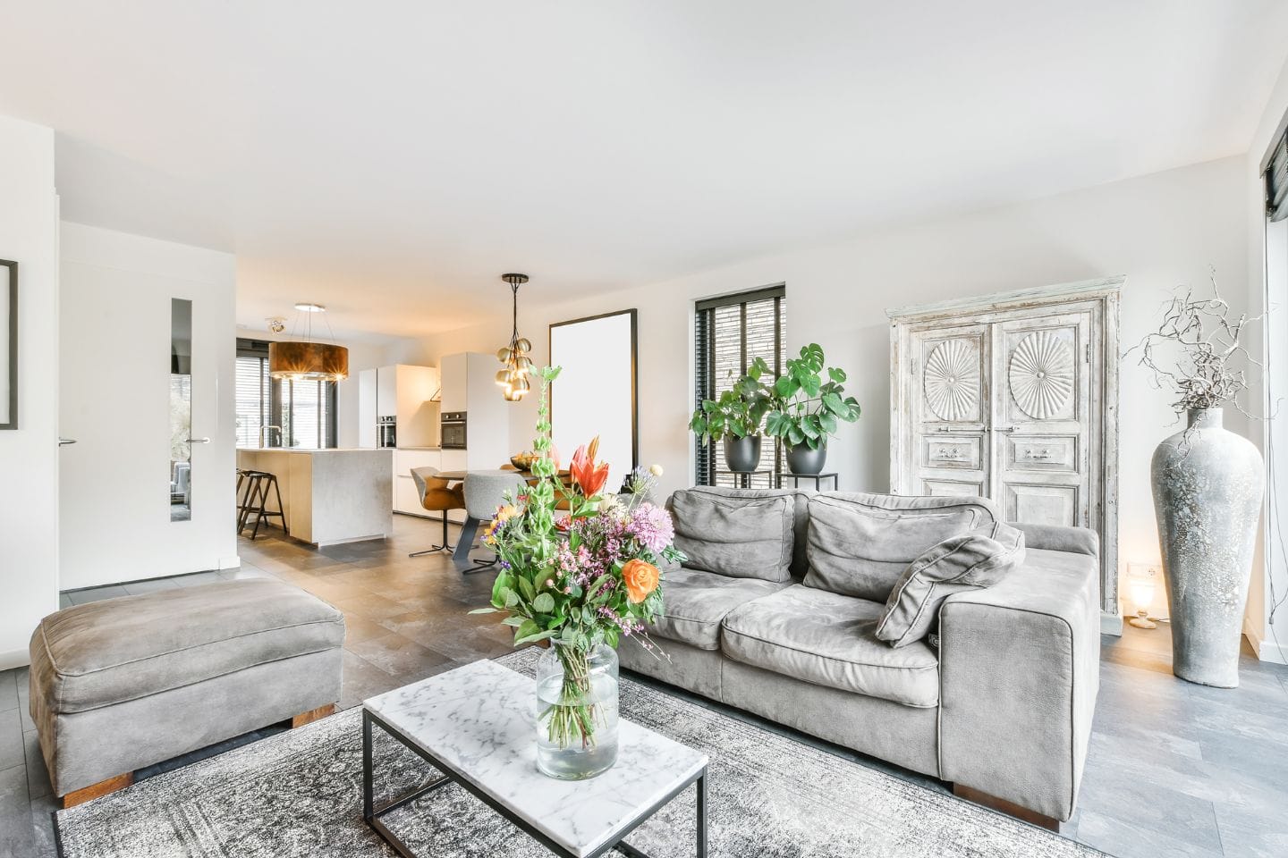 A modern, open-plan living room with a grey sofa, glass-topped coffee table with flowers, and a large potted plant. A kitchen with a bar area is visible in the background.