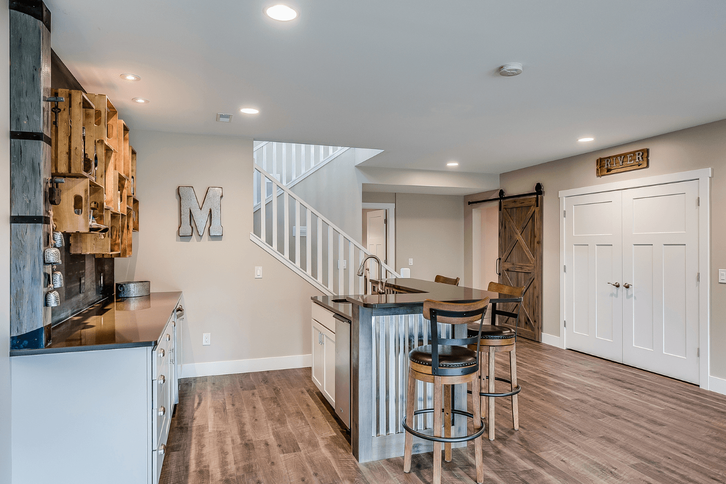 A basement kitchen with a metal-framed island, two barstools, rustic wooden cabinets, and barn-style sliding doors. There's a decorative letter "M" on the wall and a staircase leading upstairs.