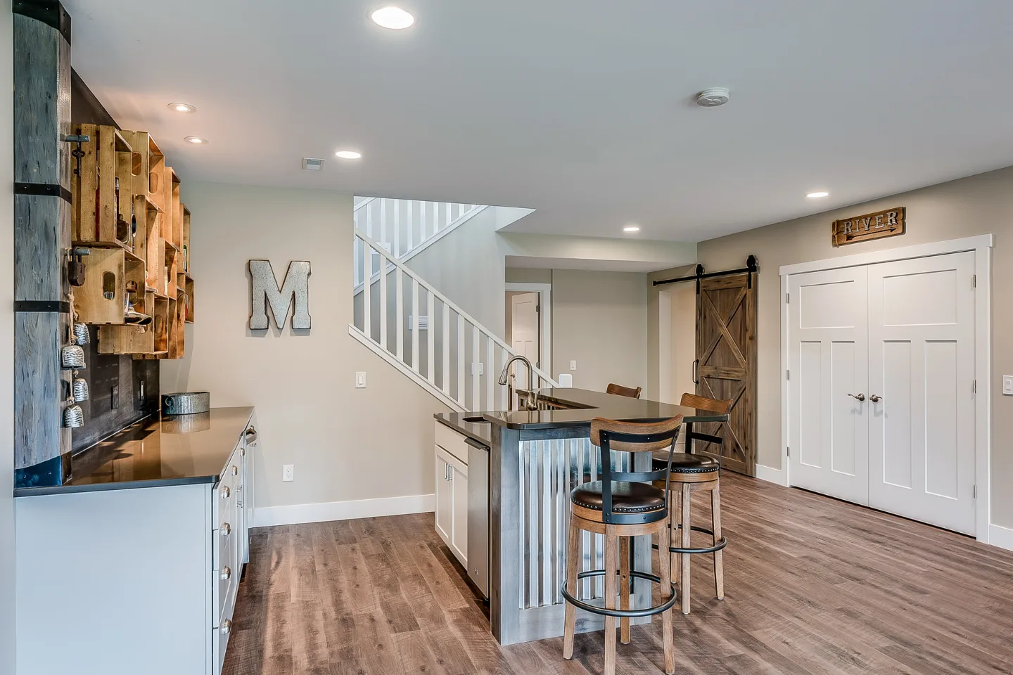 A basement kitchen with a metal-framed island, two barstools, rustic wooden cabinets, and barn-style sliding doors. There's a decorative letter "M" on the wall and a staircase leading upstairs.