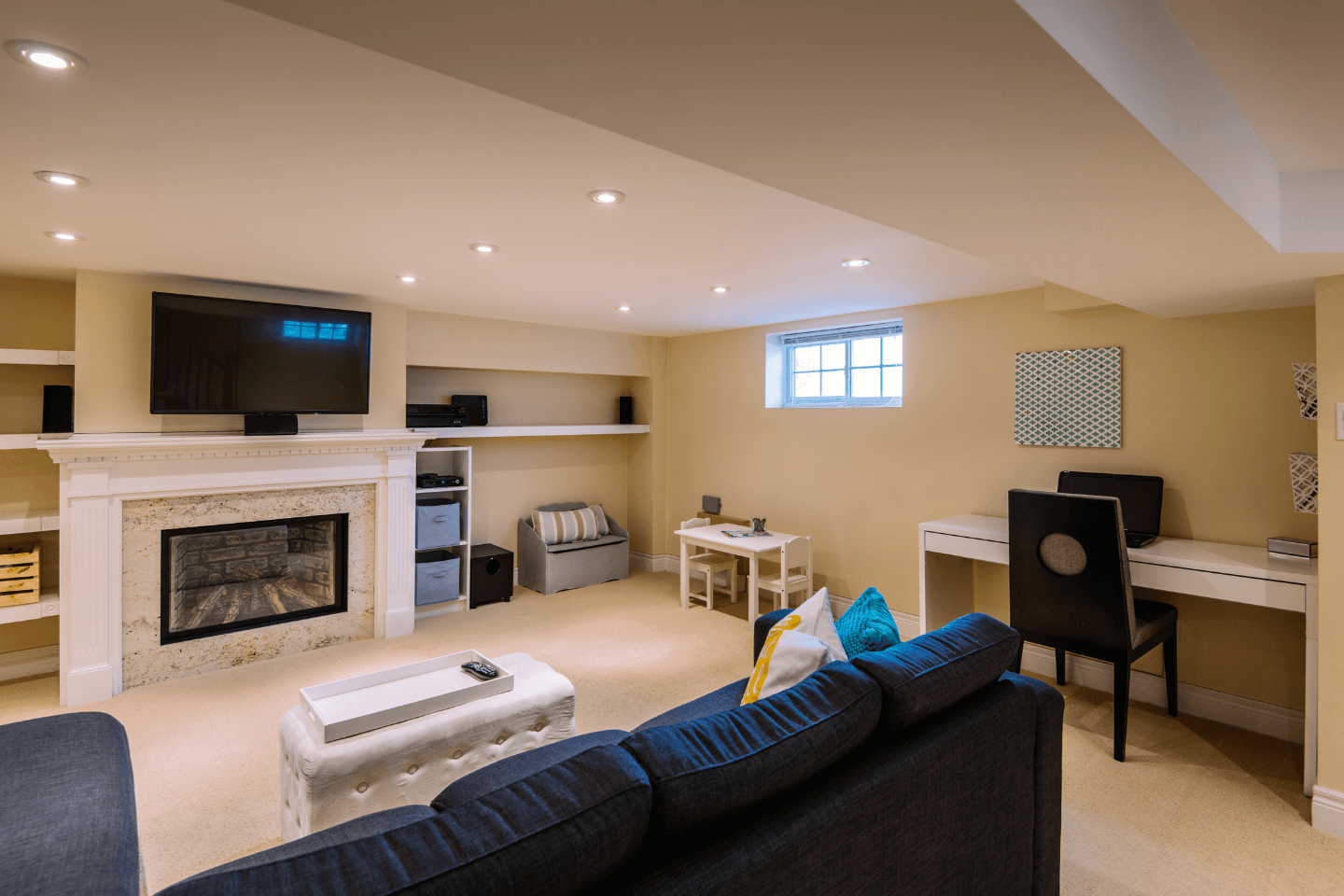 A basement living area with a fireplace, wall-mounted TV, desk, and seating including a couch and armchair. The room has beige walls, carpeted flooring, and recessed lighting.