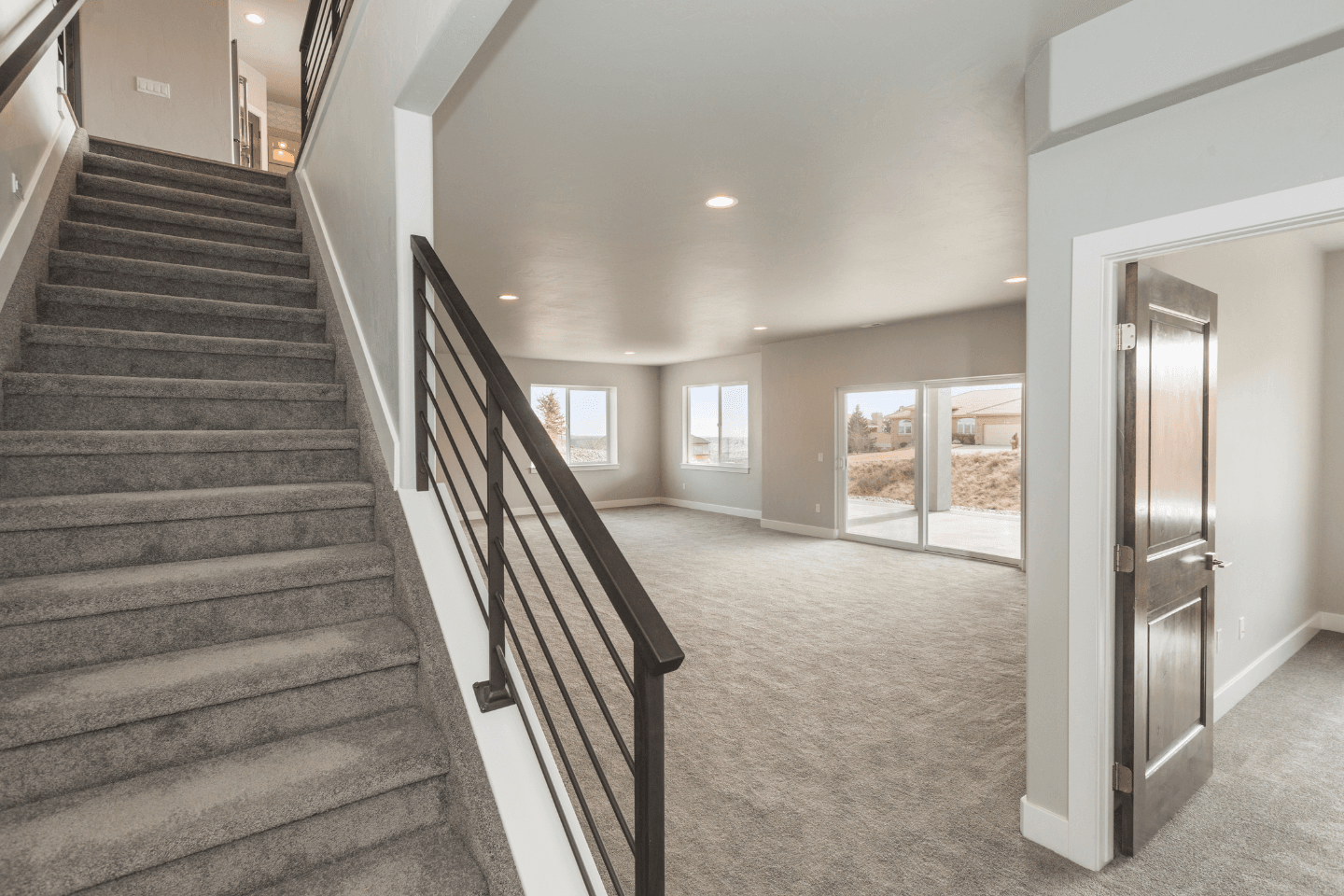 A modern, carpeted living area with large windows, a staircase on the left with a black handrail, and an open doorway on the right. Natural light illuminates the room.