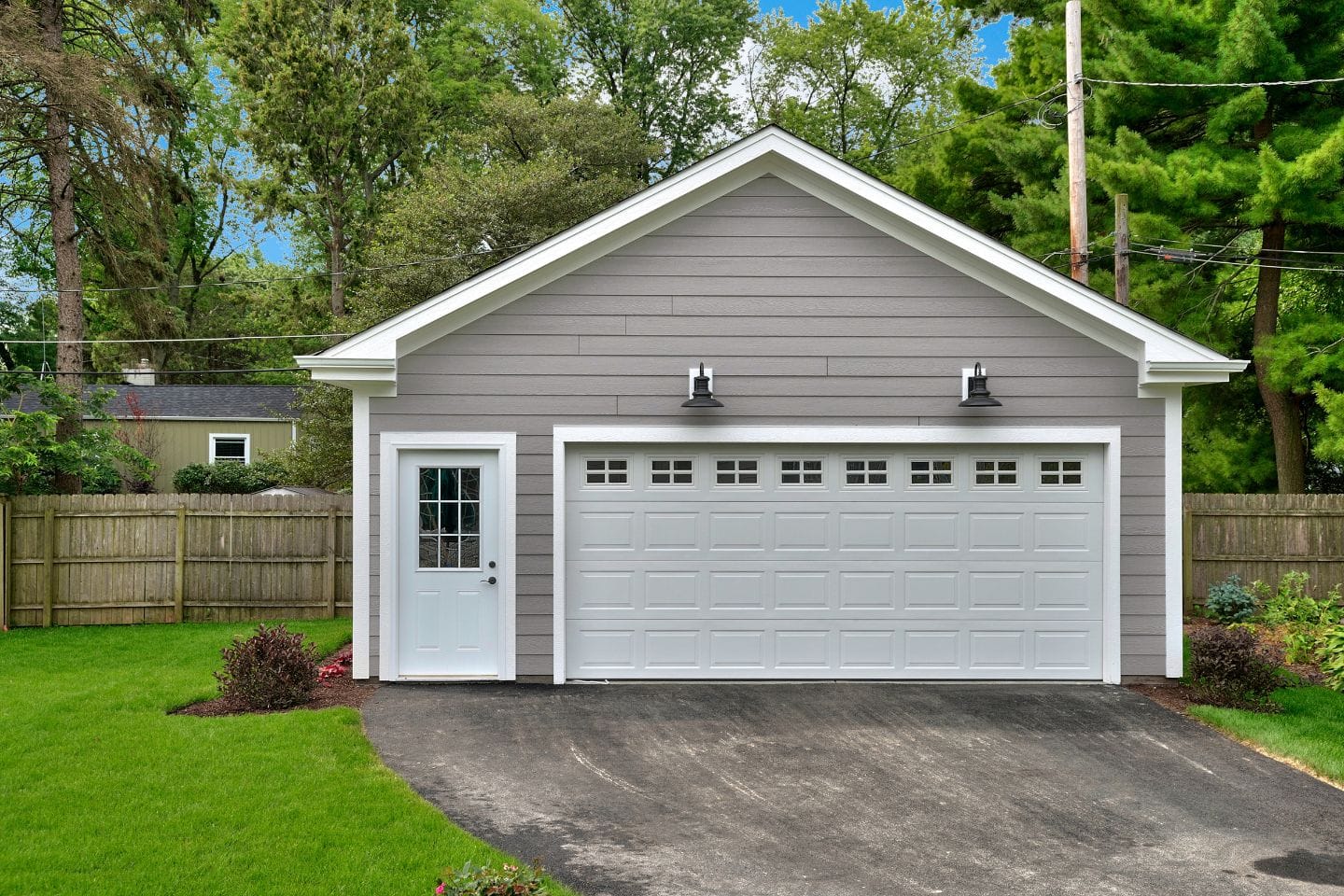 A grey detached garage with a white door and windows, surrounded by greenery and a wooden fence, offers potential for conversion into an ADU or secondary unit.