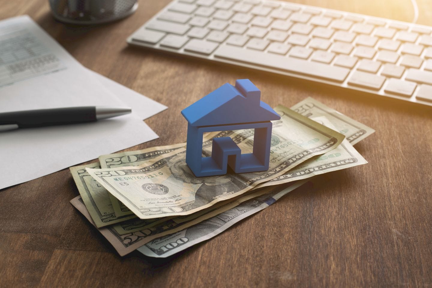 A small blue house model is placed on top of scattered US dollar bills on a wooden desk, alongside a keyboard, pen, and documents—an ideal setup attracting savvy investors.