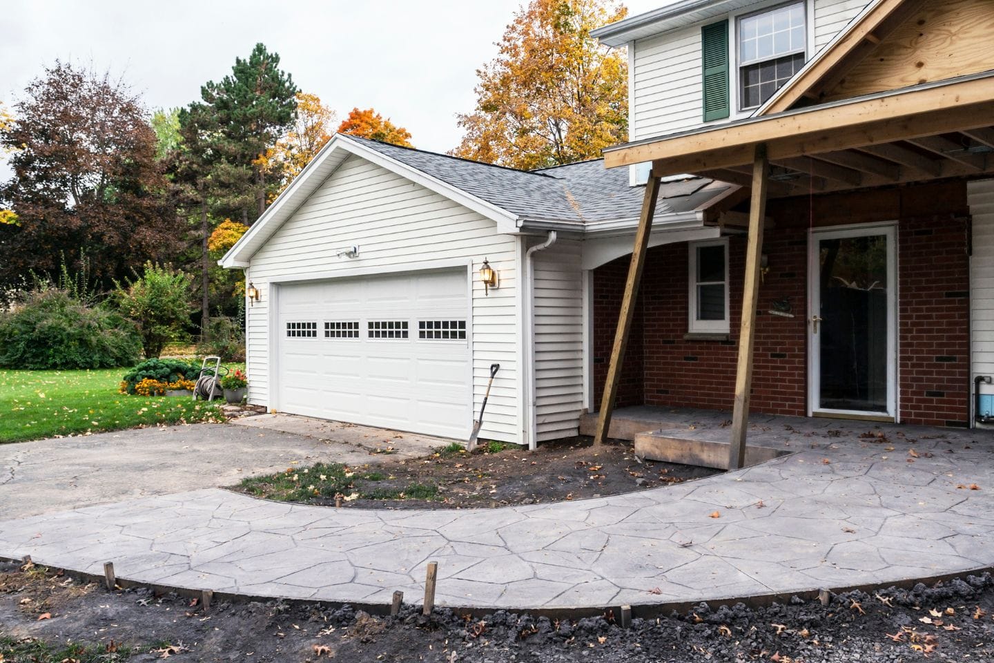 A grey concrete walkway in front of a garage and house under construction with wooden framework seen by the entrance; grass, trees, and autumn foliage in the background reflects ongoing home renovations.