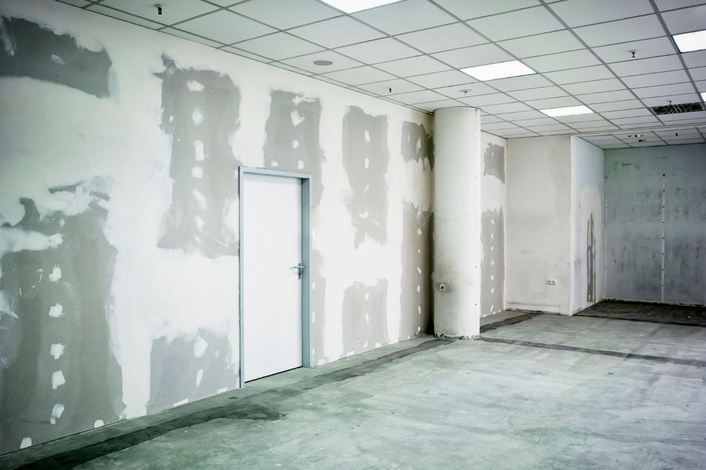 An unfinished room with partially completed drywall, a closed door, and a column. The ceiling features fluorescent lights and exposed areas. The bare concrete floor speaks to the potential of this space, ideal for home additions or house renovations.