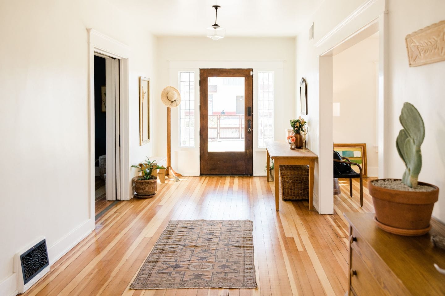 A well-lit hallway with wooden flooring, a rug, plants, a wooden console with decor, and a door at the far end. Two rooms are visible on either side, showcasing thoughtful home renovations that enhance both style and function.